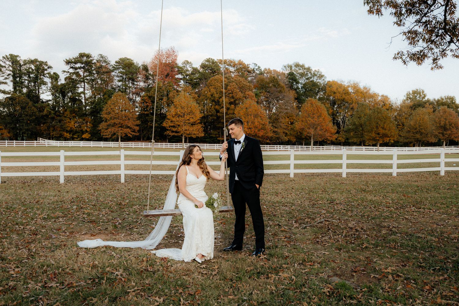 A couple in wedding attire stands together on an autumn day in front of a white fence at a rustic farm setting.