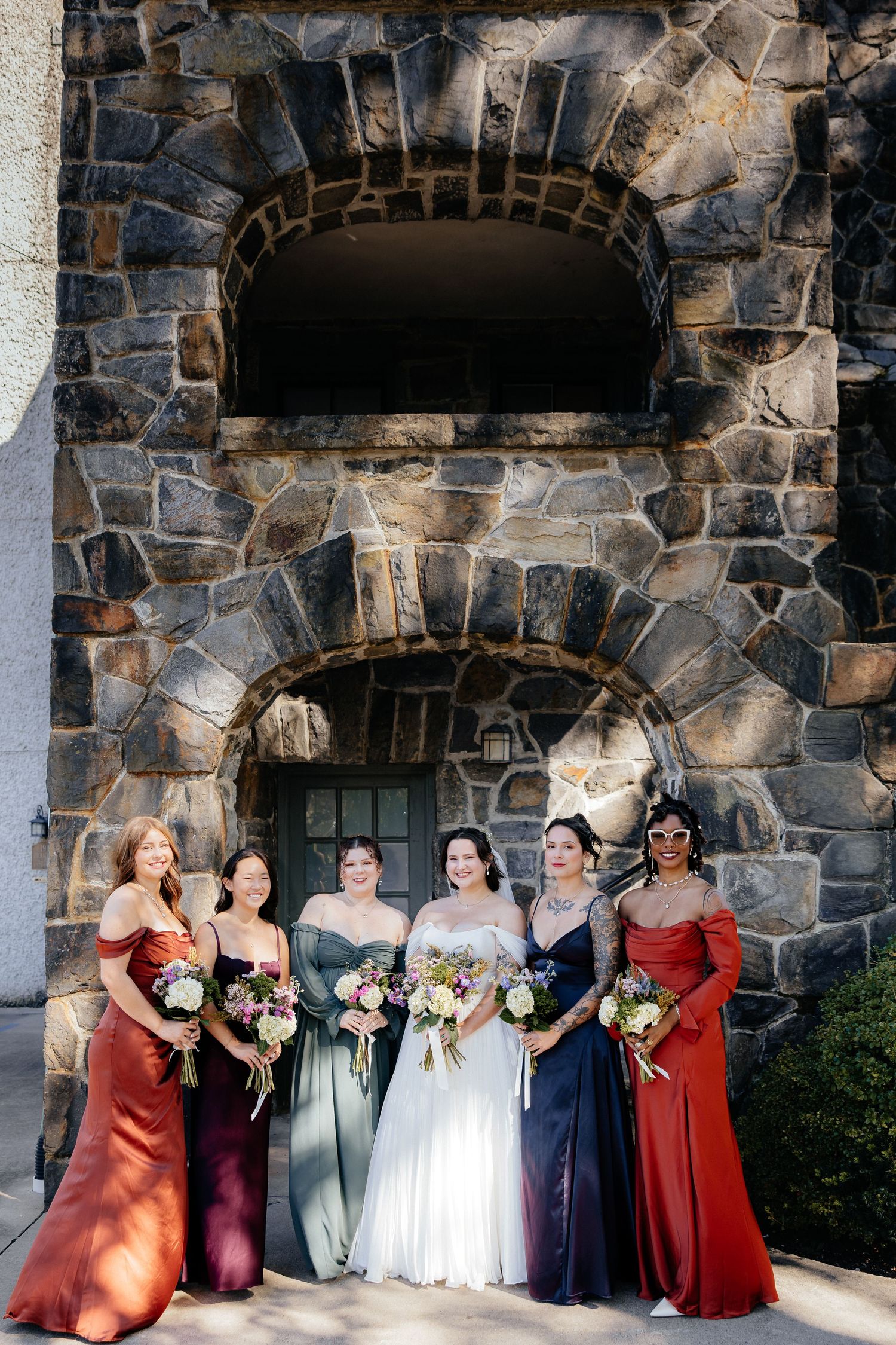 Bride in a white gown stands with four bridesmaids in rust, plum, sage and navy dresses holding bouquets before a stone archway.
