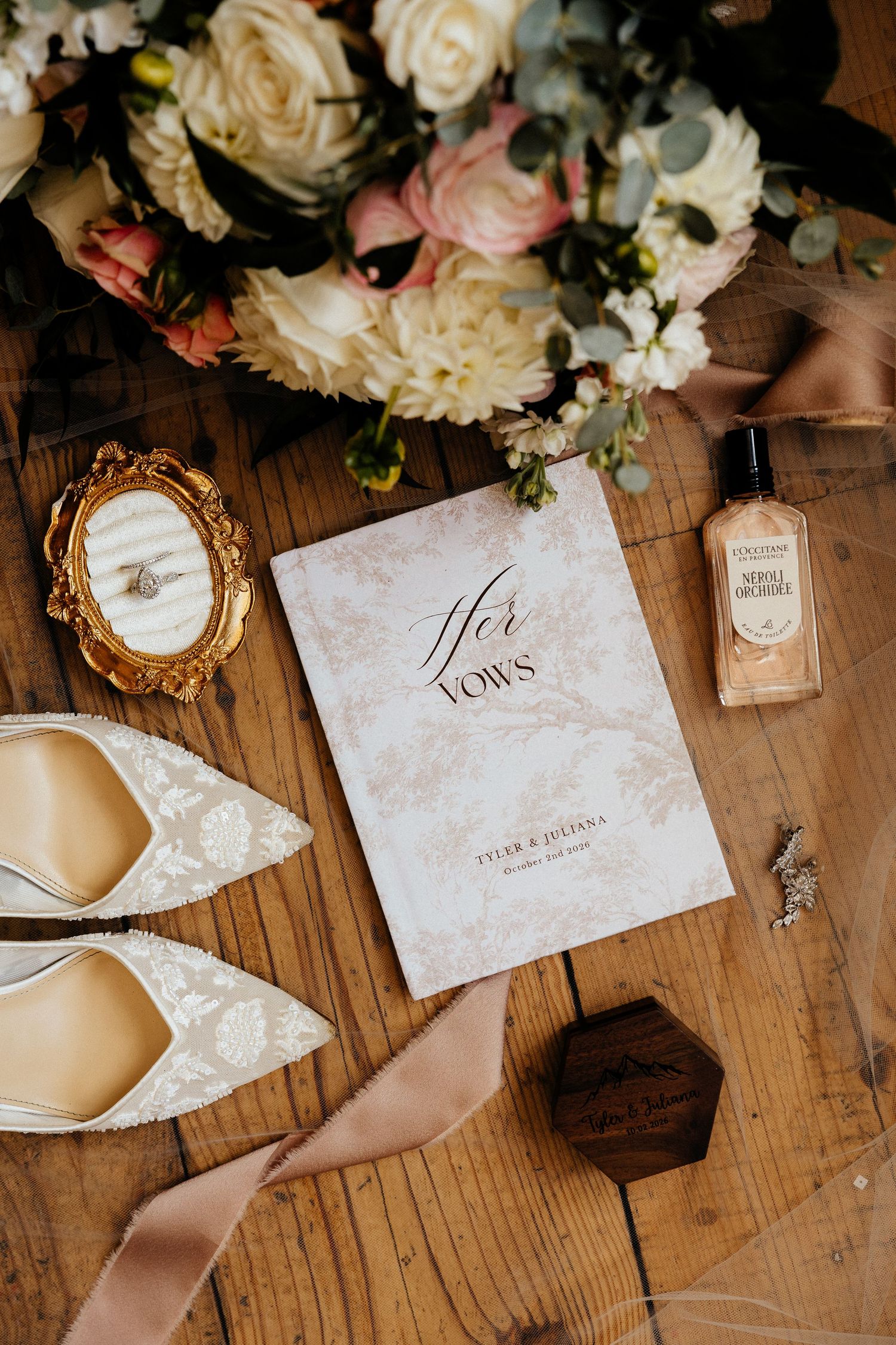 Wedding flat lay with a Her Vows booklet, lace bridal heels, a cream rose bouquet, perfume bottle, and ring box on rustic wood.
