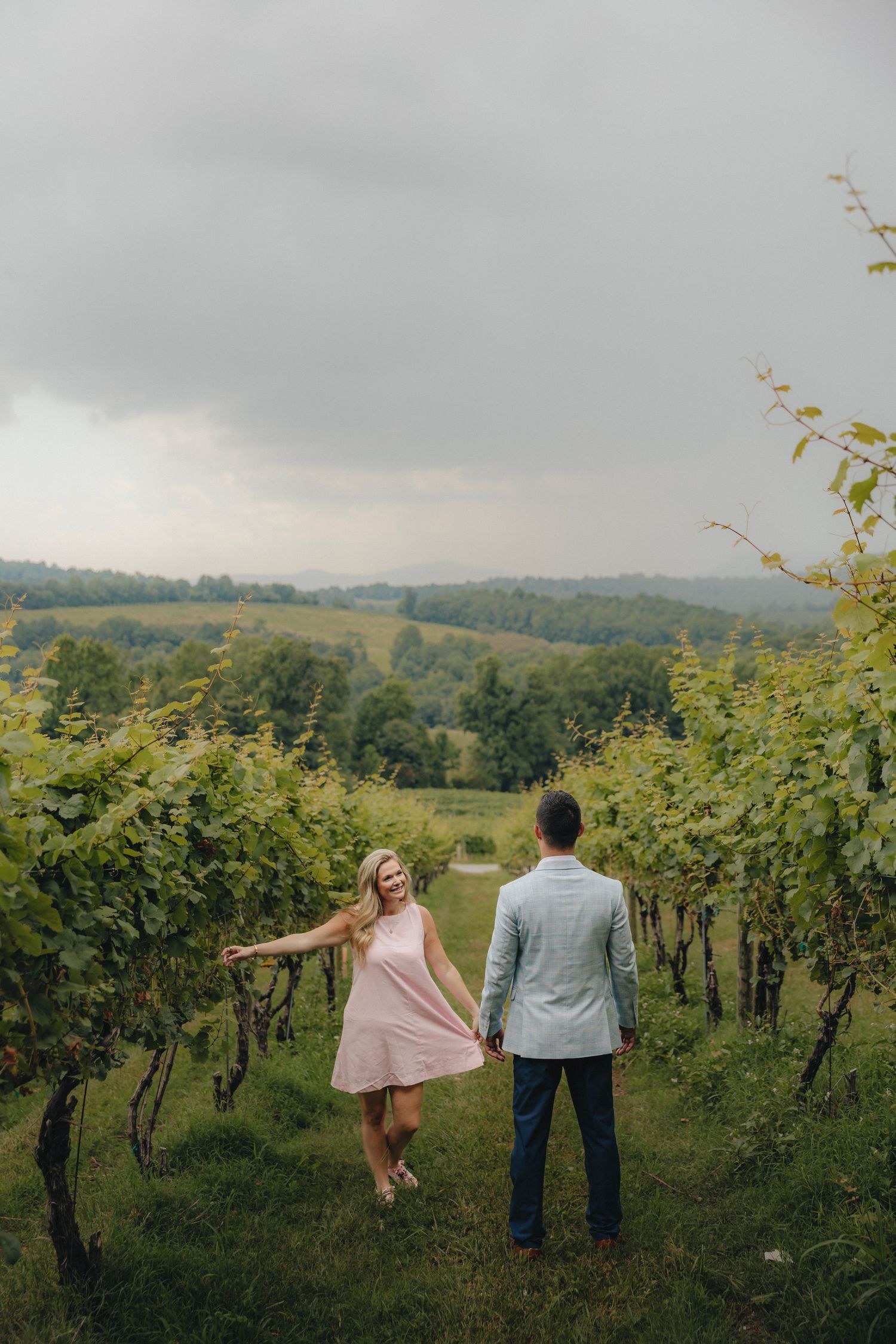 Couple holding hands in a lush vineyard, woman in pink dress smiling, rolling hills in background.