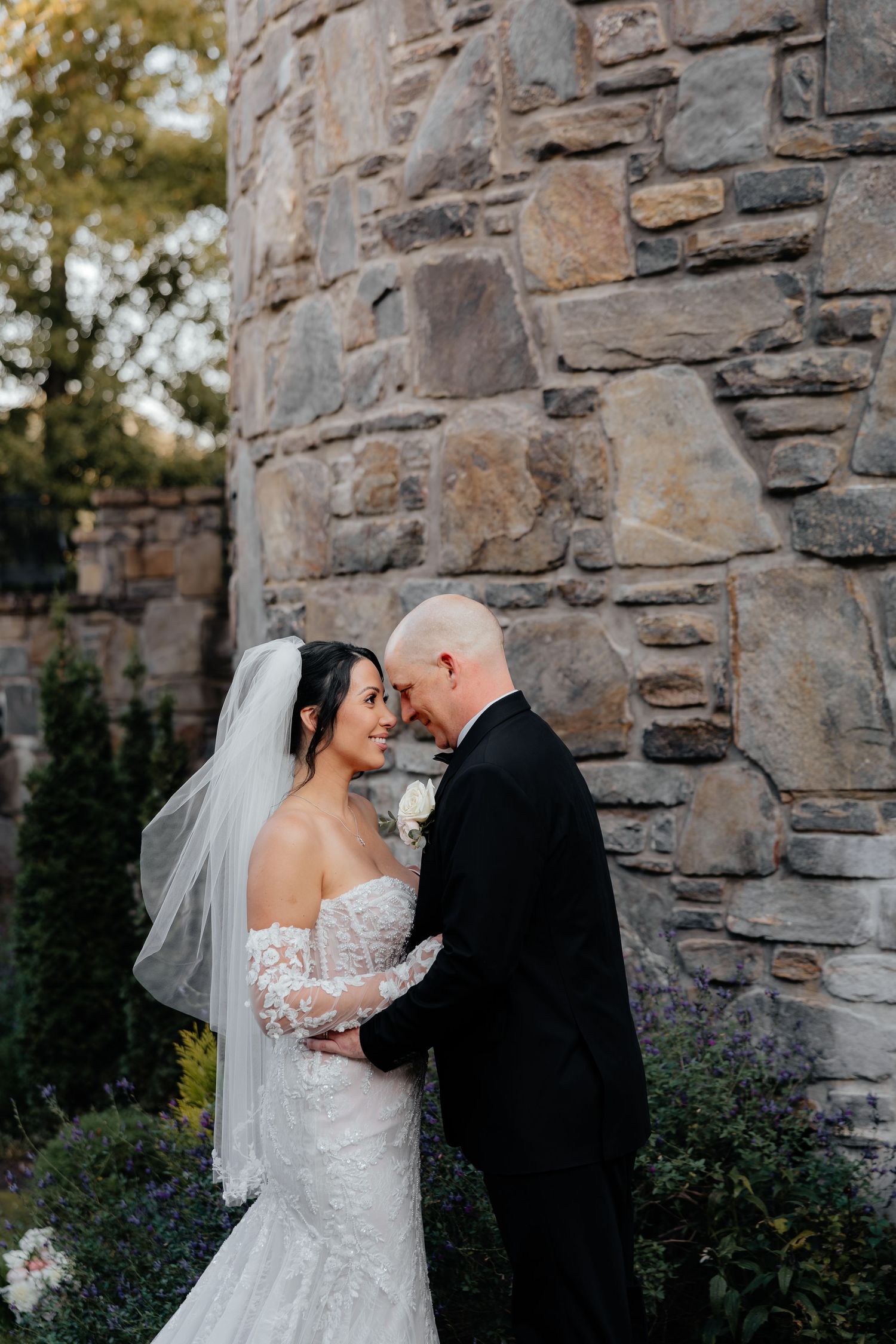 Bride in a lace wedding gown and veil shares an intimate moment with her groom in a black suit beside a rustic stone wall.