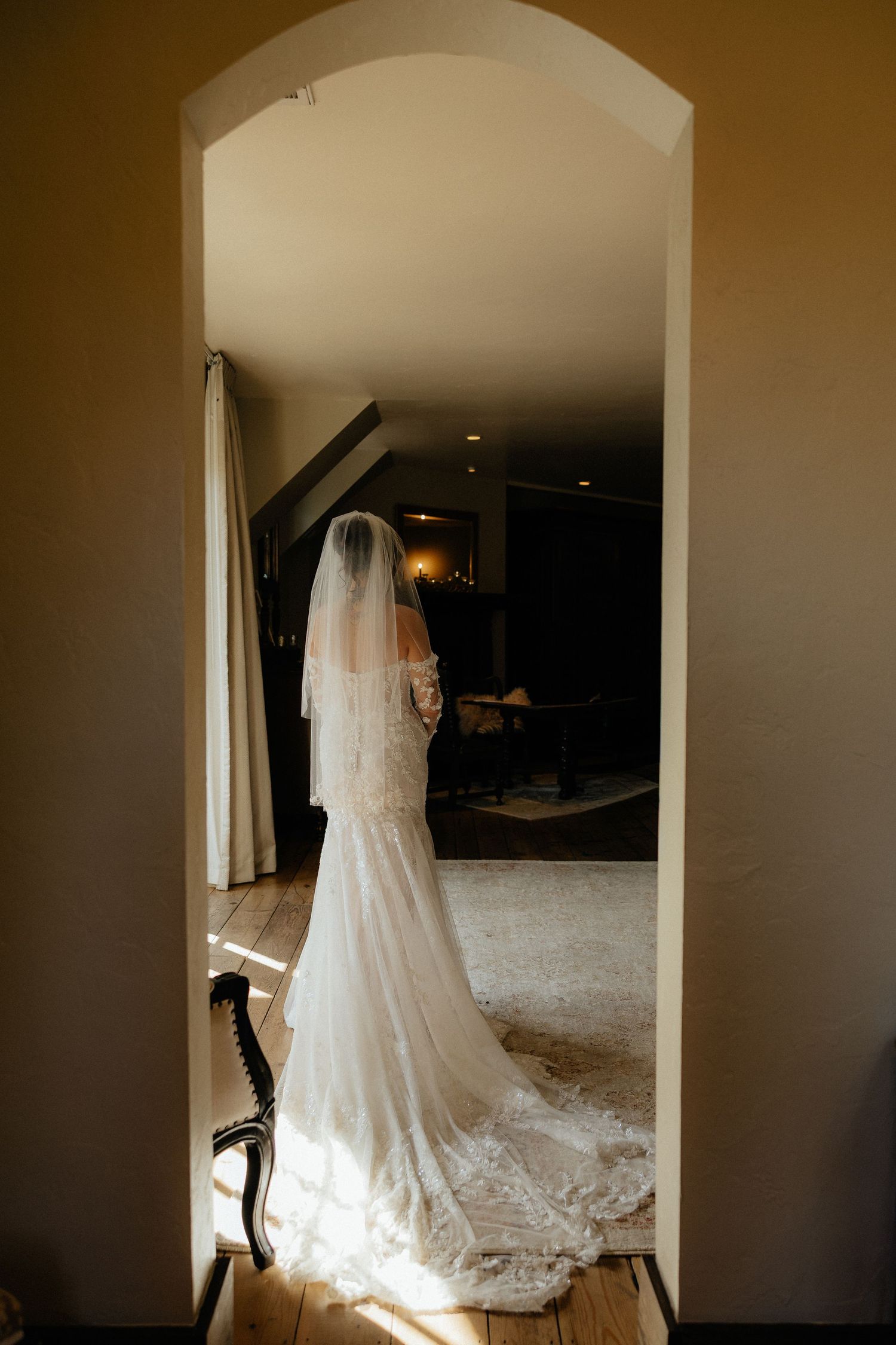 Bride in lace wedding gown and long veil stands in sunlit doorway, elegant train flowing on hardwood floor.