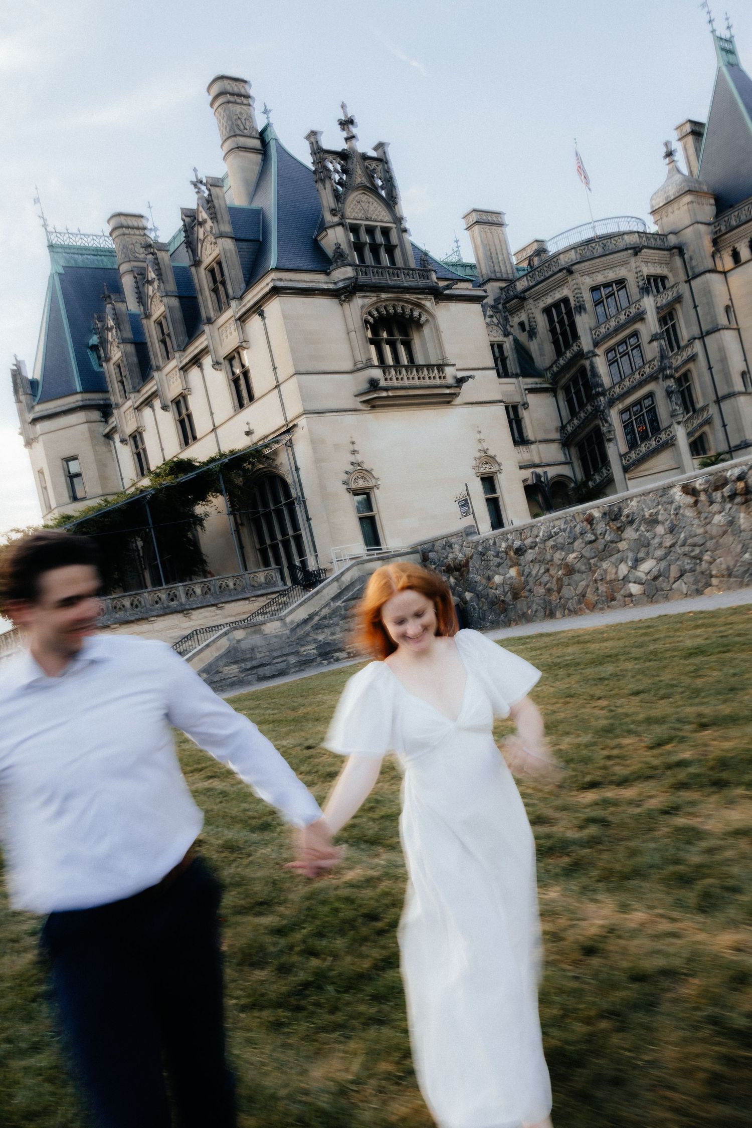 Bride in white dress and groom run hand-in-hand laughing in front of grand Biltmore Estate.
