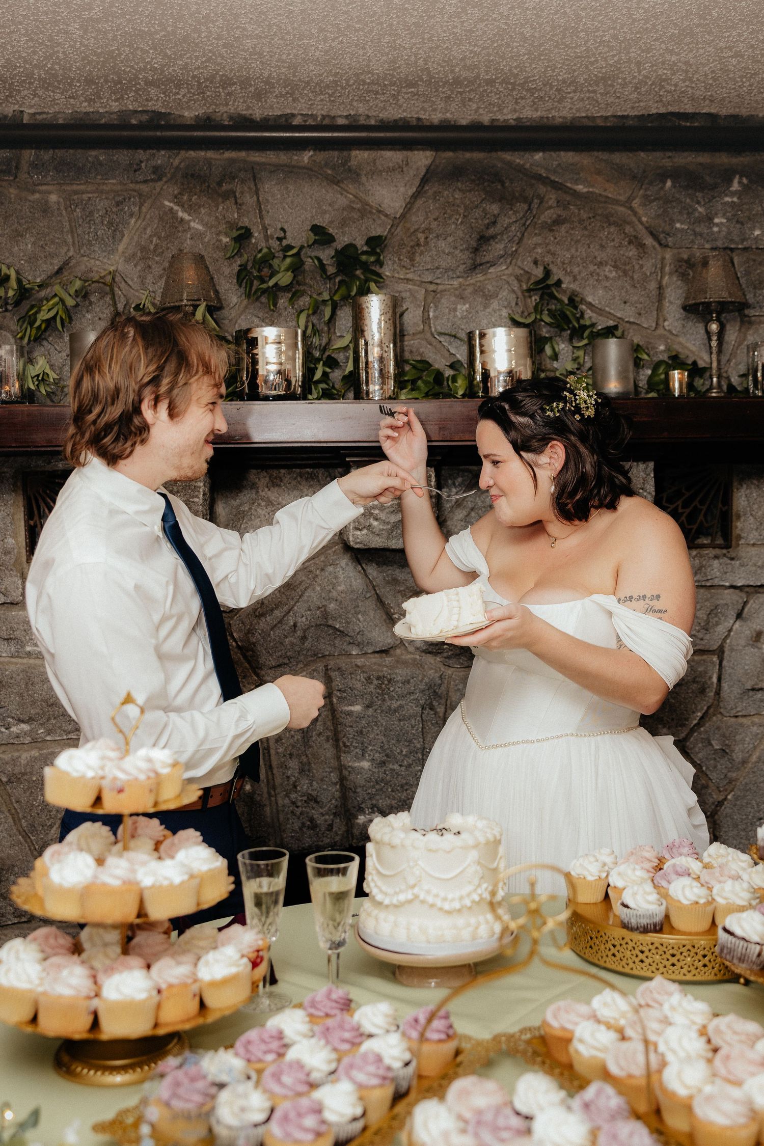 Bride and groom playfully feed each other wedding cake at a dessert table filled with frosted cupcakes and a small white cake.