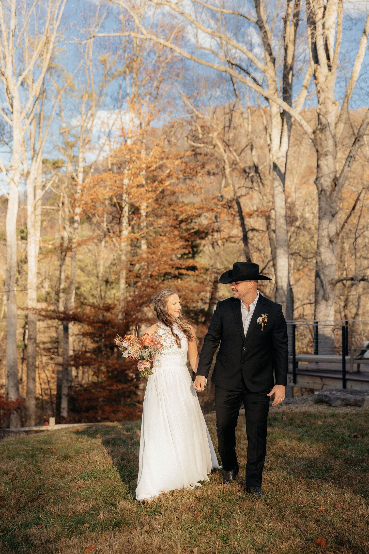 A couple stands together outdoors during an autumn wedding with bare trees and fall foliage in the background.