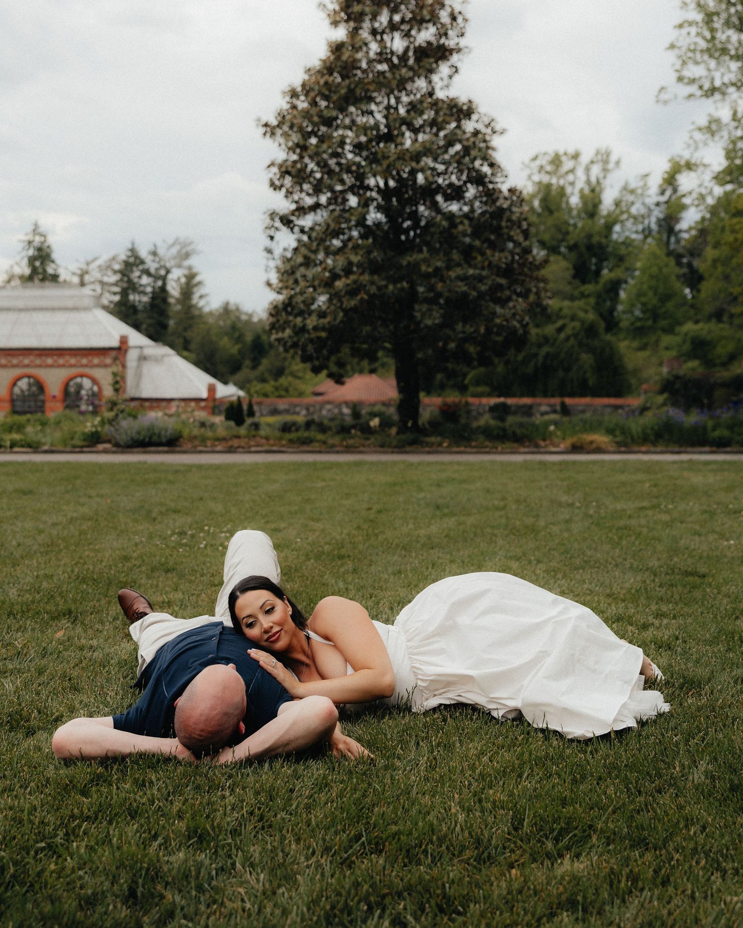 Bride and groom laughing together on green grass at outdoor wedding venue with brick building backdrop.