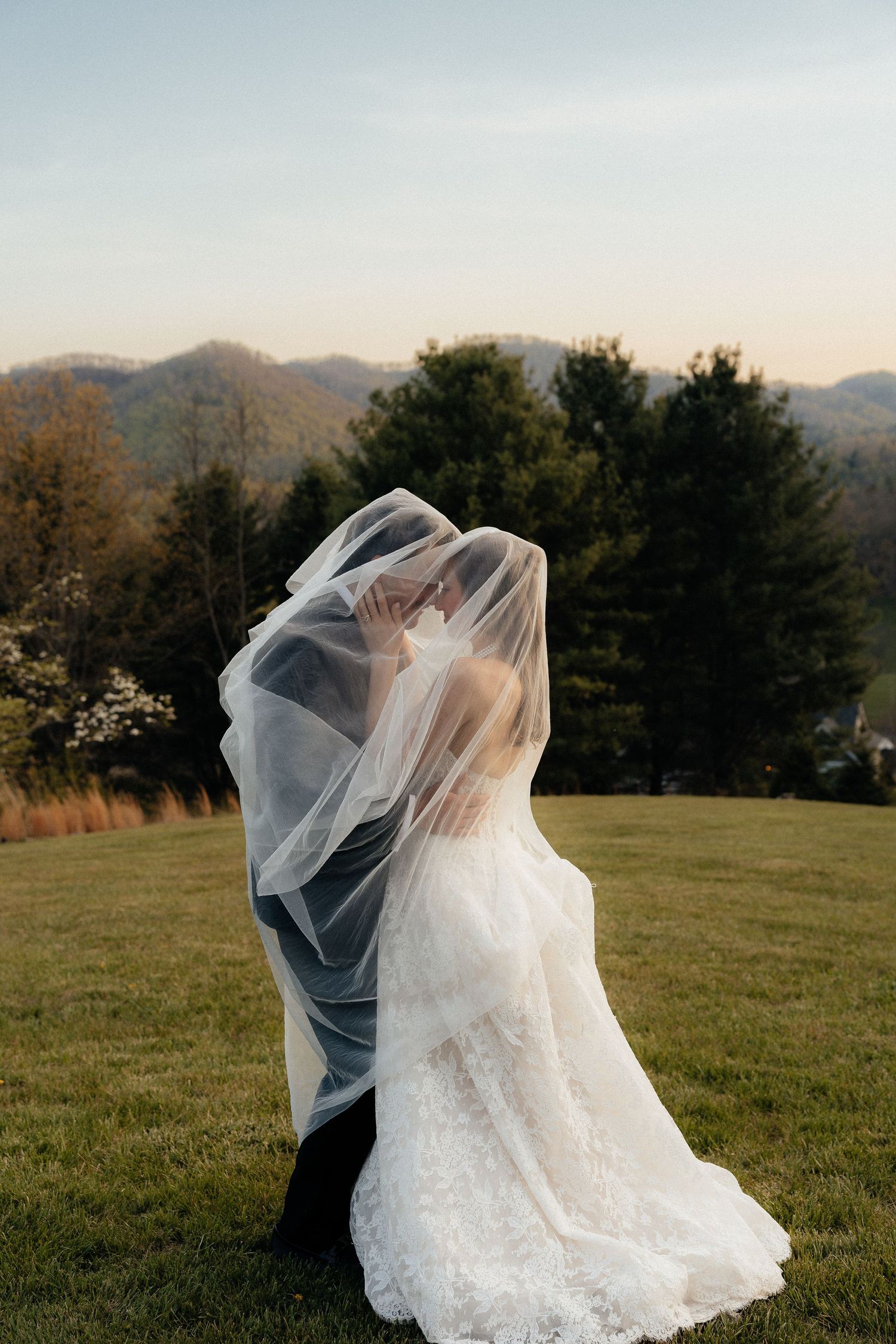 Bride and groom embrace under a sheer wedding veil on a grassy hilltop with evergreen trees and mountains in the background at sunset.