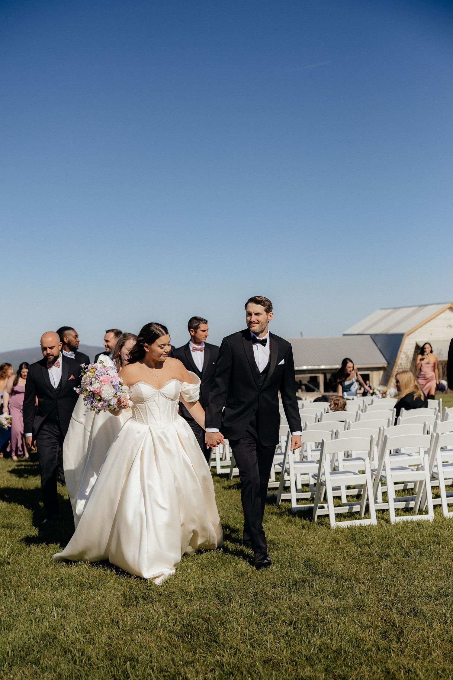 Bride and groom walk hand-in-hand down the aisle after outdoor wedding ceremony on a sunny day.