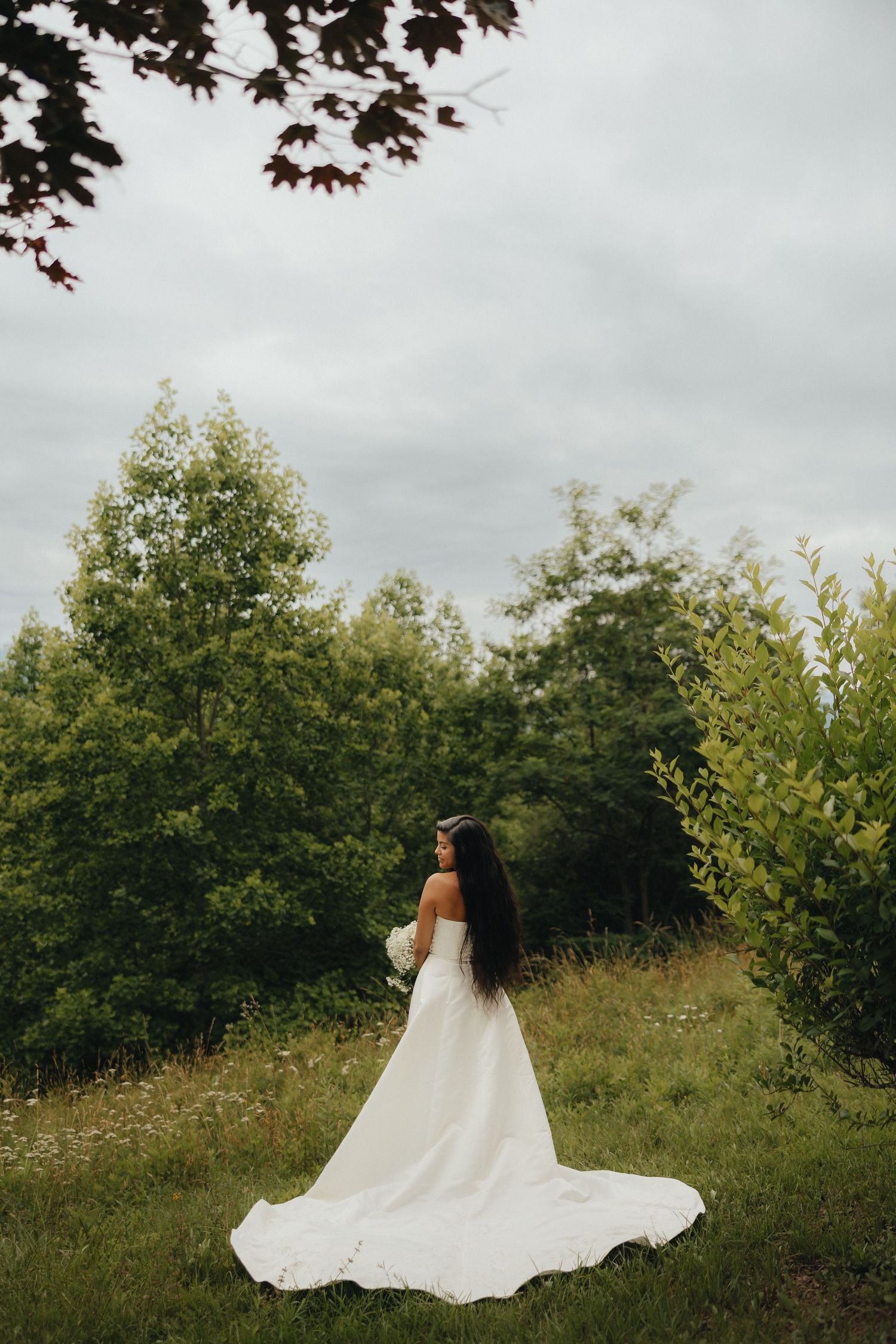 Bride in white off-shoulder gown with long train stands in lush green forest clearing on overcast day.