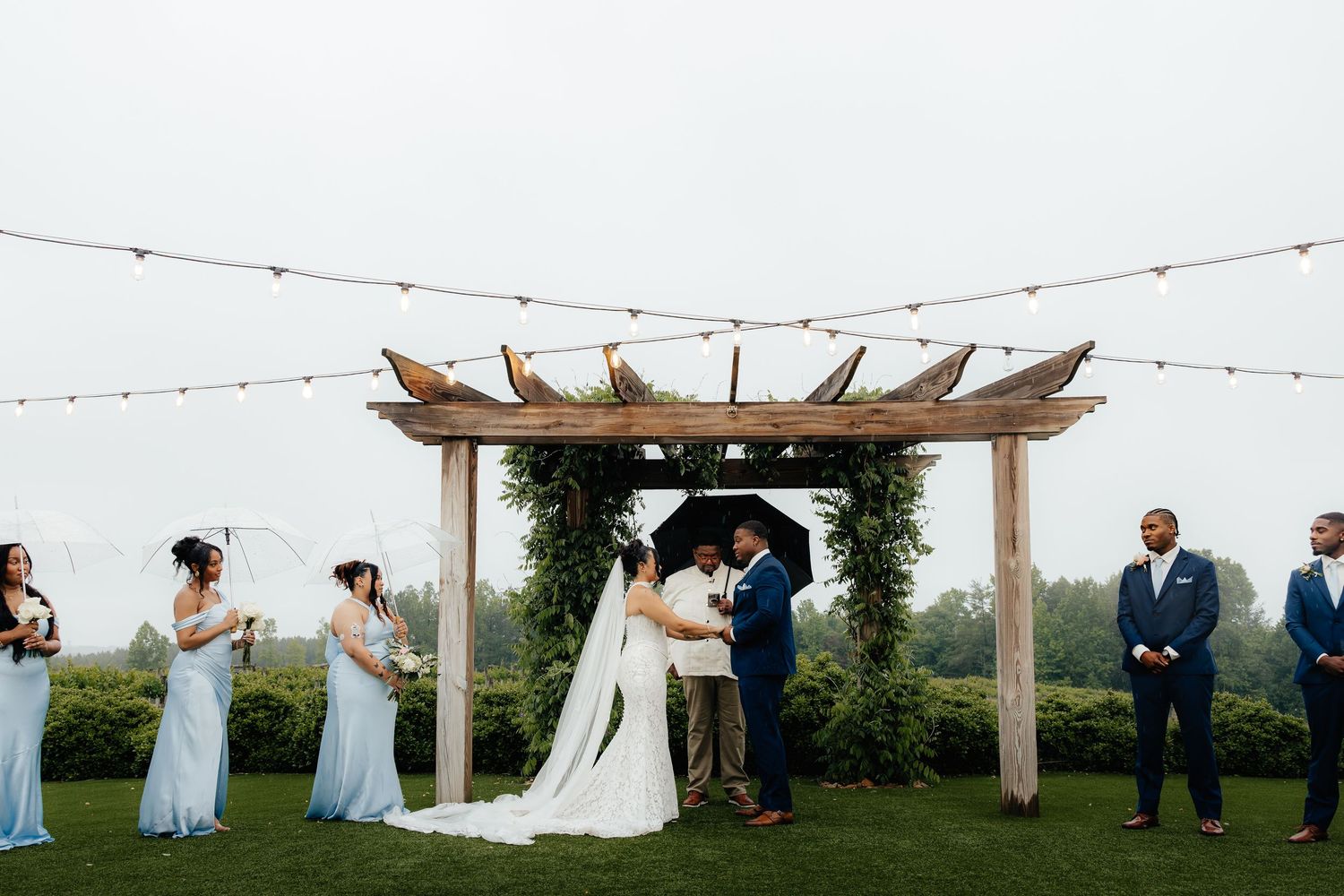 Bride and groom share a first kiss under a wooden pergola arch with string lights during an outdoor wedding ceremony.