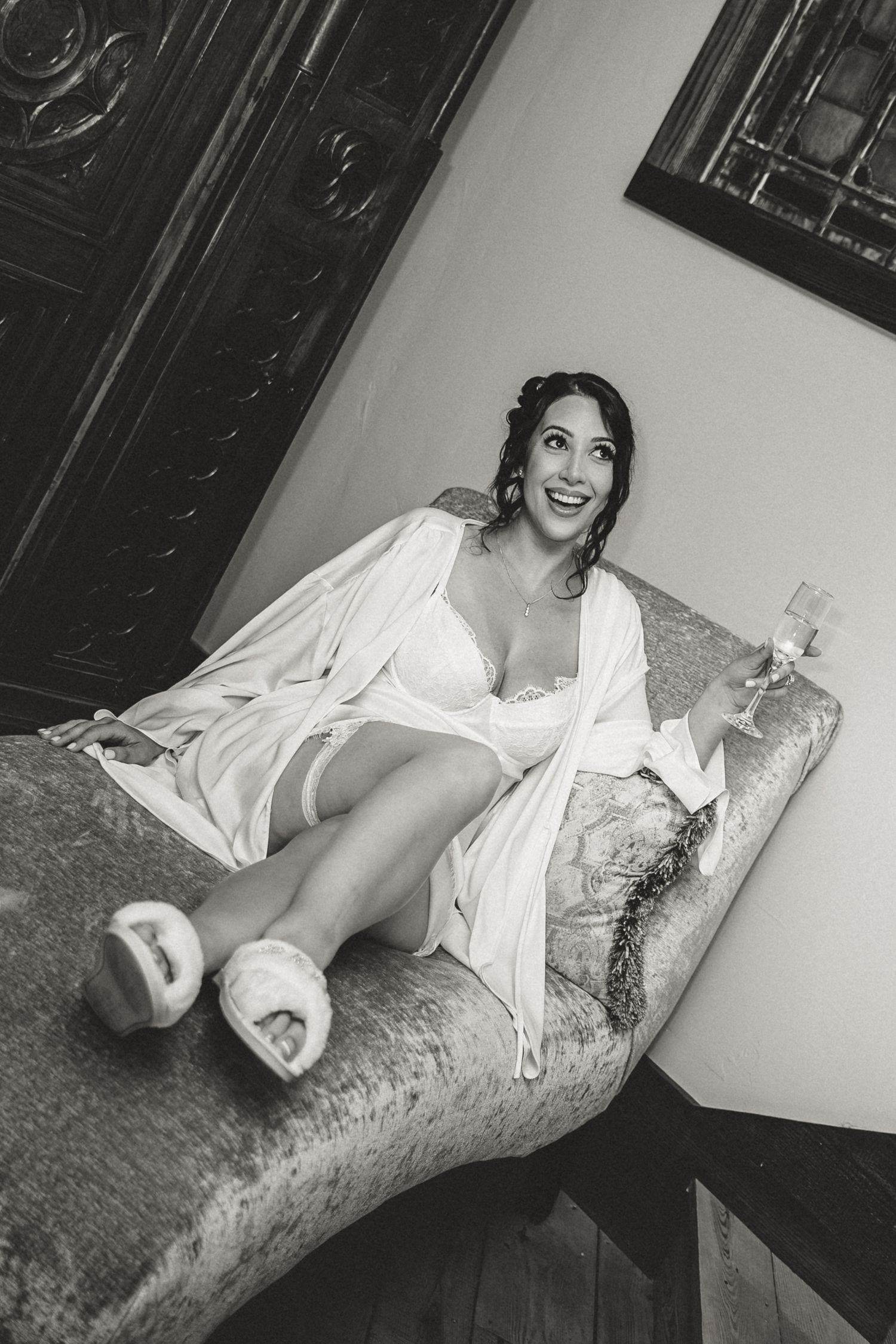 Black and white bridal boudoir photo of a laughing bride in a white robe and fuzzy slippers holding champagne on a vintage chaise.