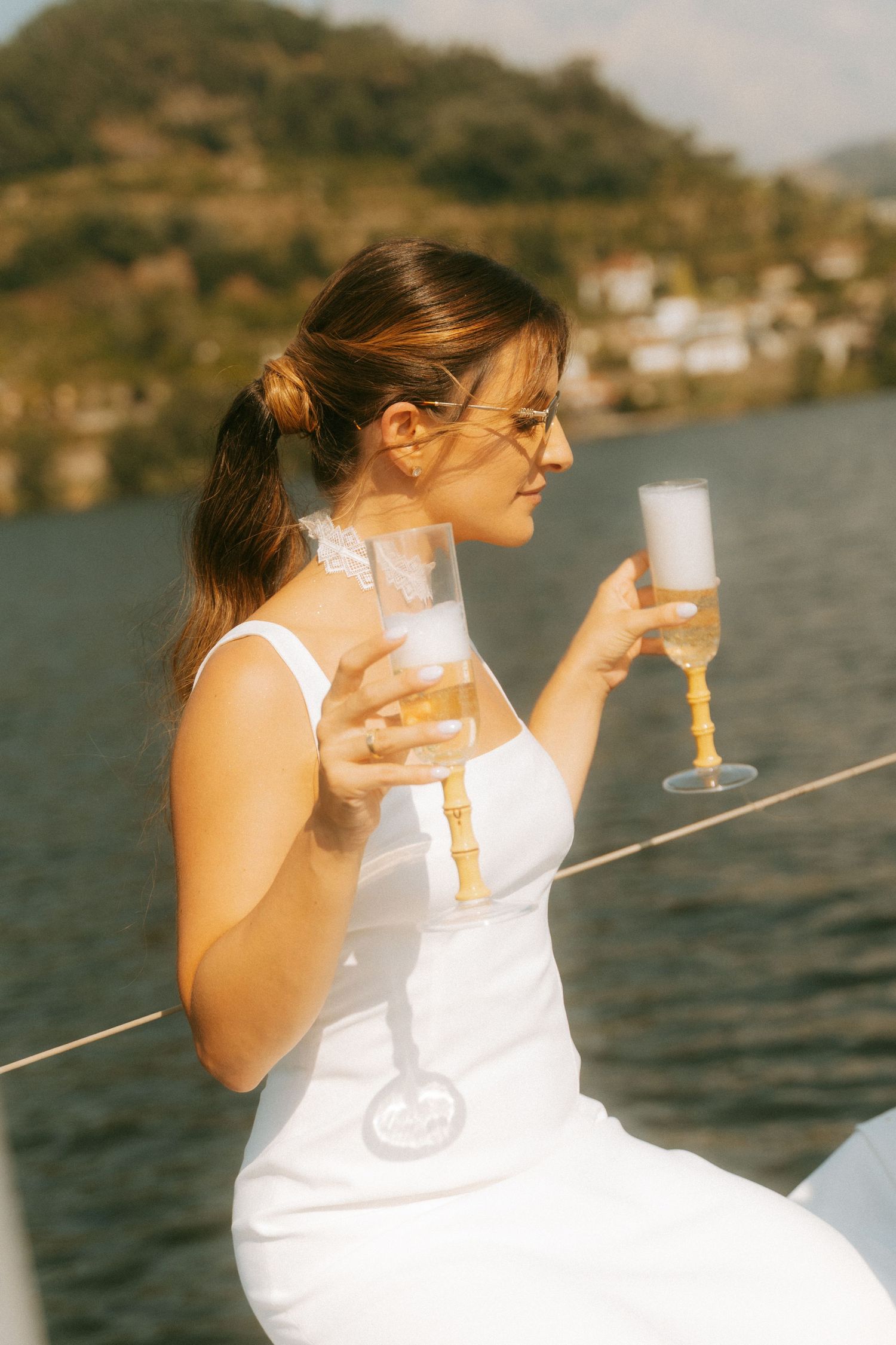 Bride in white dress holding champagne flutes on a sailboat with scenic waterfront village backdrop.