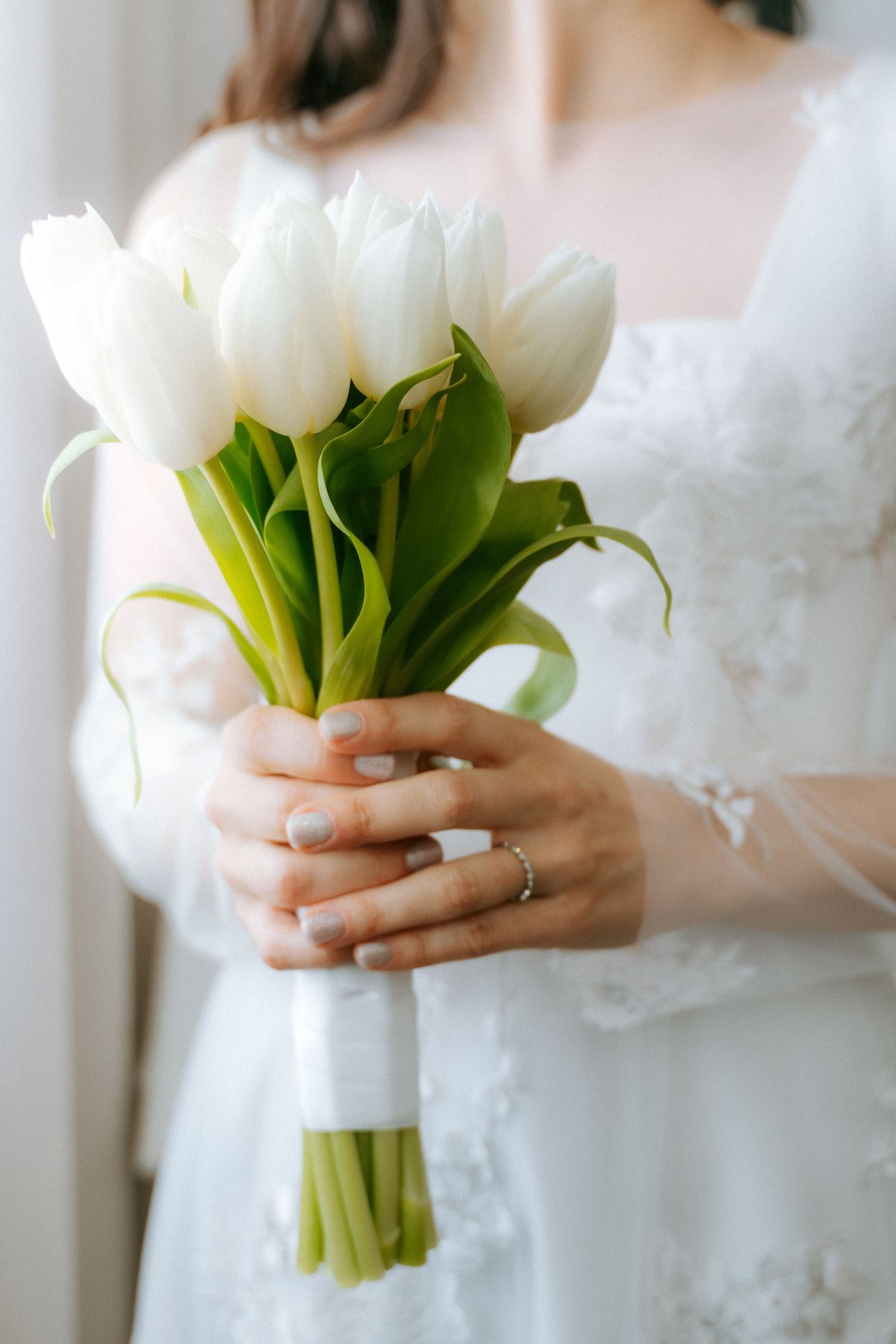Bride in lace wedding dress holding a bouquet of fresh white tulips tied with a white ribbon.