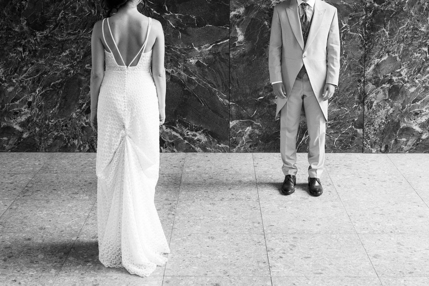Black and white photo of a bride in a backless white gown and groom in a suit standing apart on a marble floor.