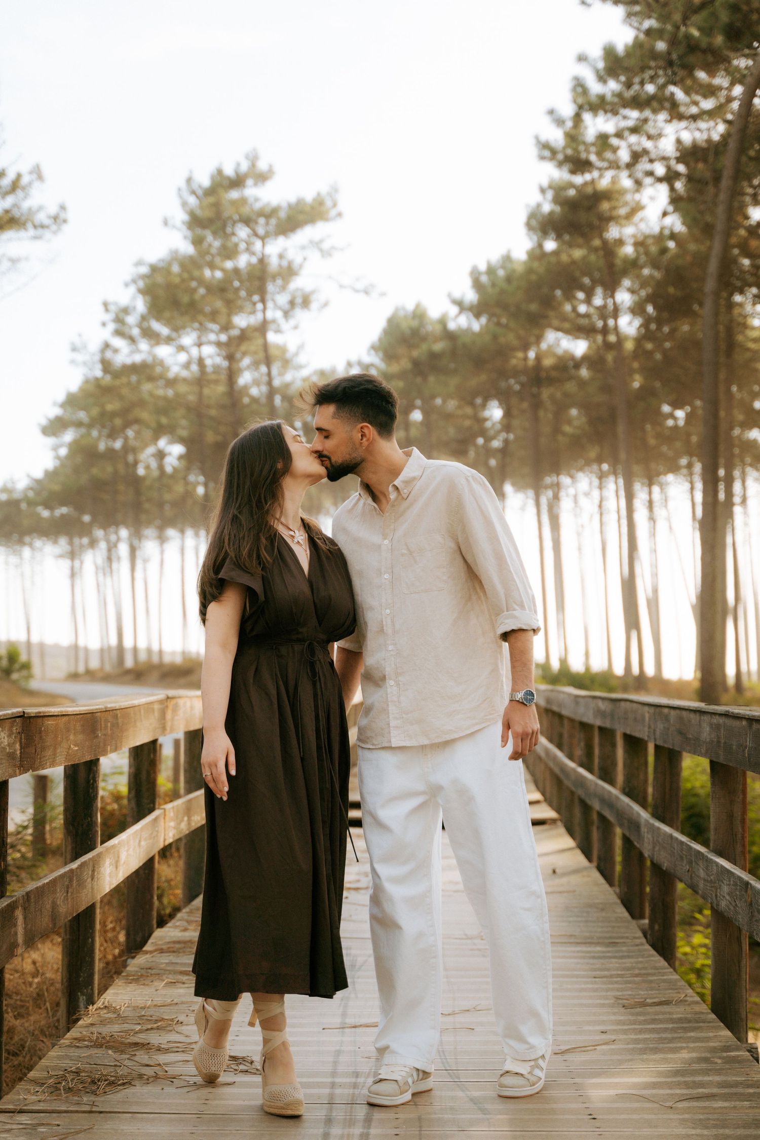 Couple sharing a romantic kiss on a wooden boardwalk surrounded by tall pine trees at sunset.