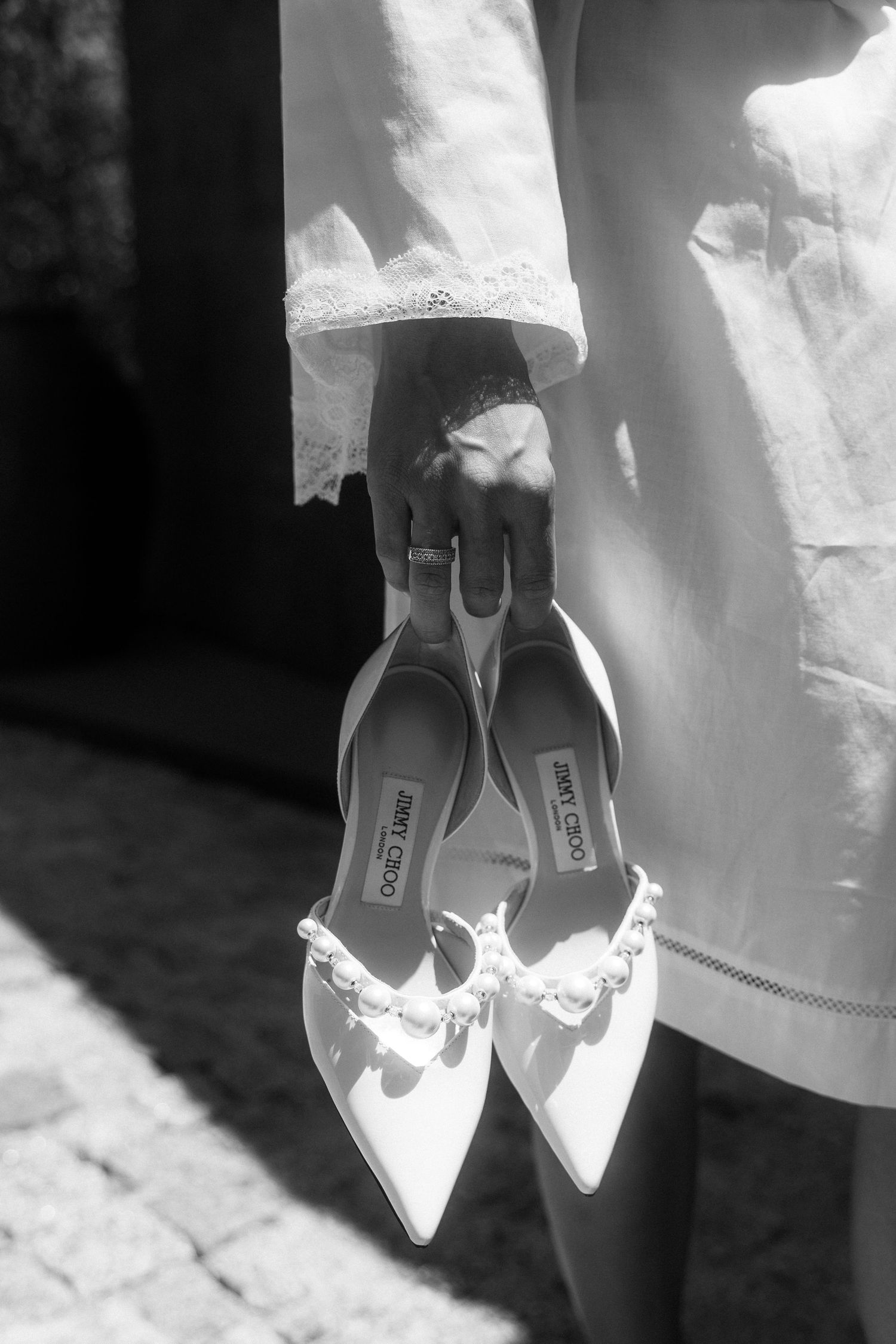 Black and white image of bride holding Jimmy Choo pearl-adorned wedding heels outdoors.