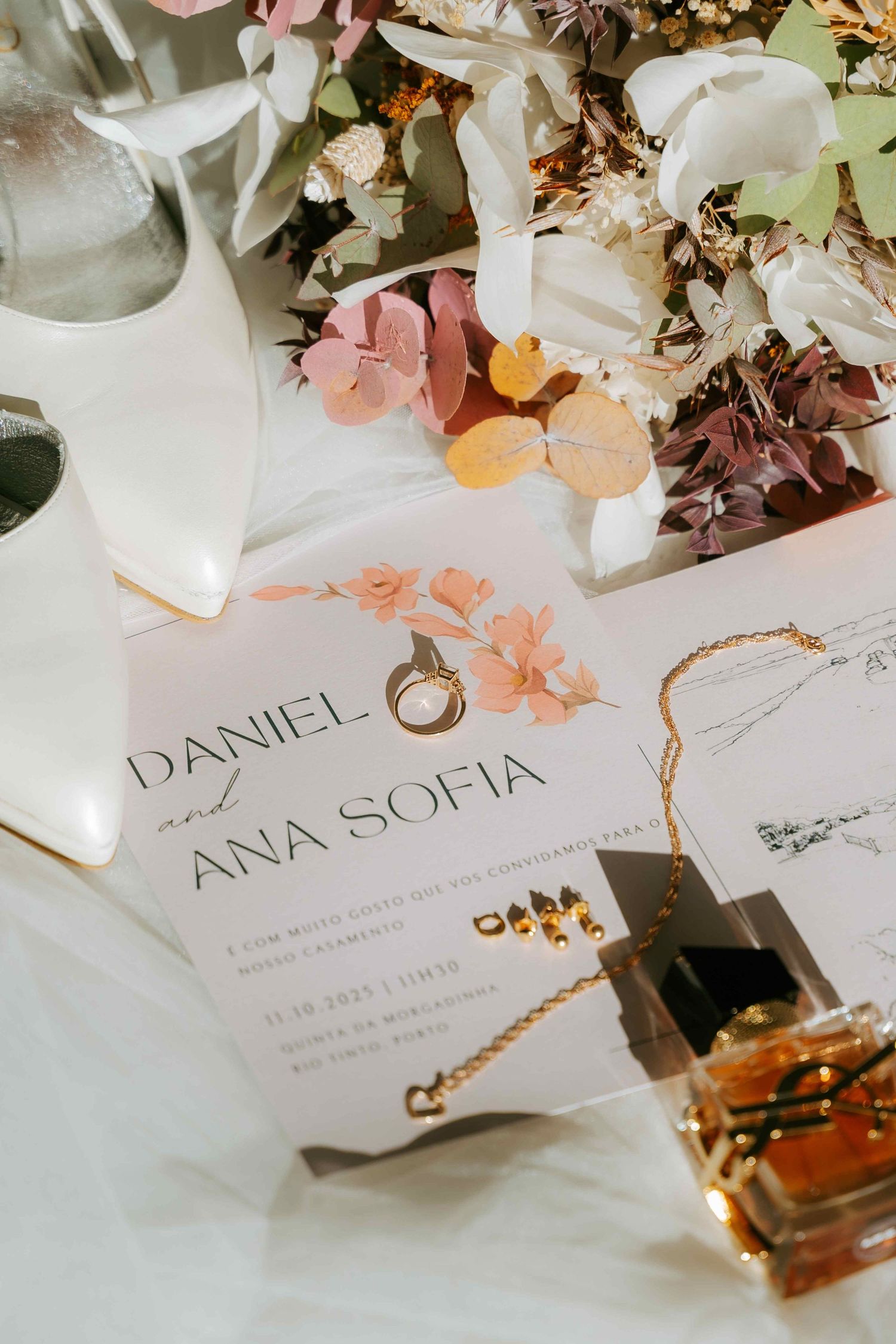 Wedding flat lay featuring Daniel and Ana Sofia invitation with floral details, rings, and perfume.