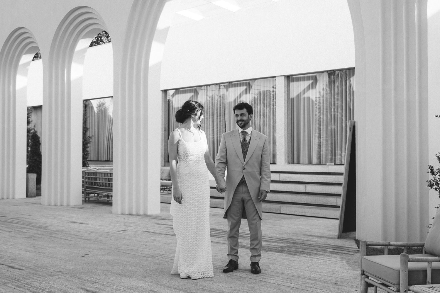 Bride and groom stand apart holding hands between elegant white columns in black and white photo.