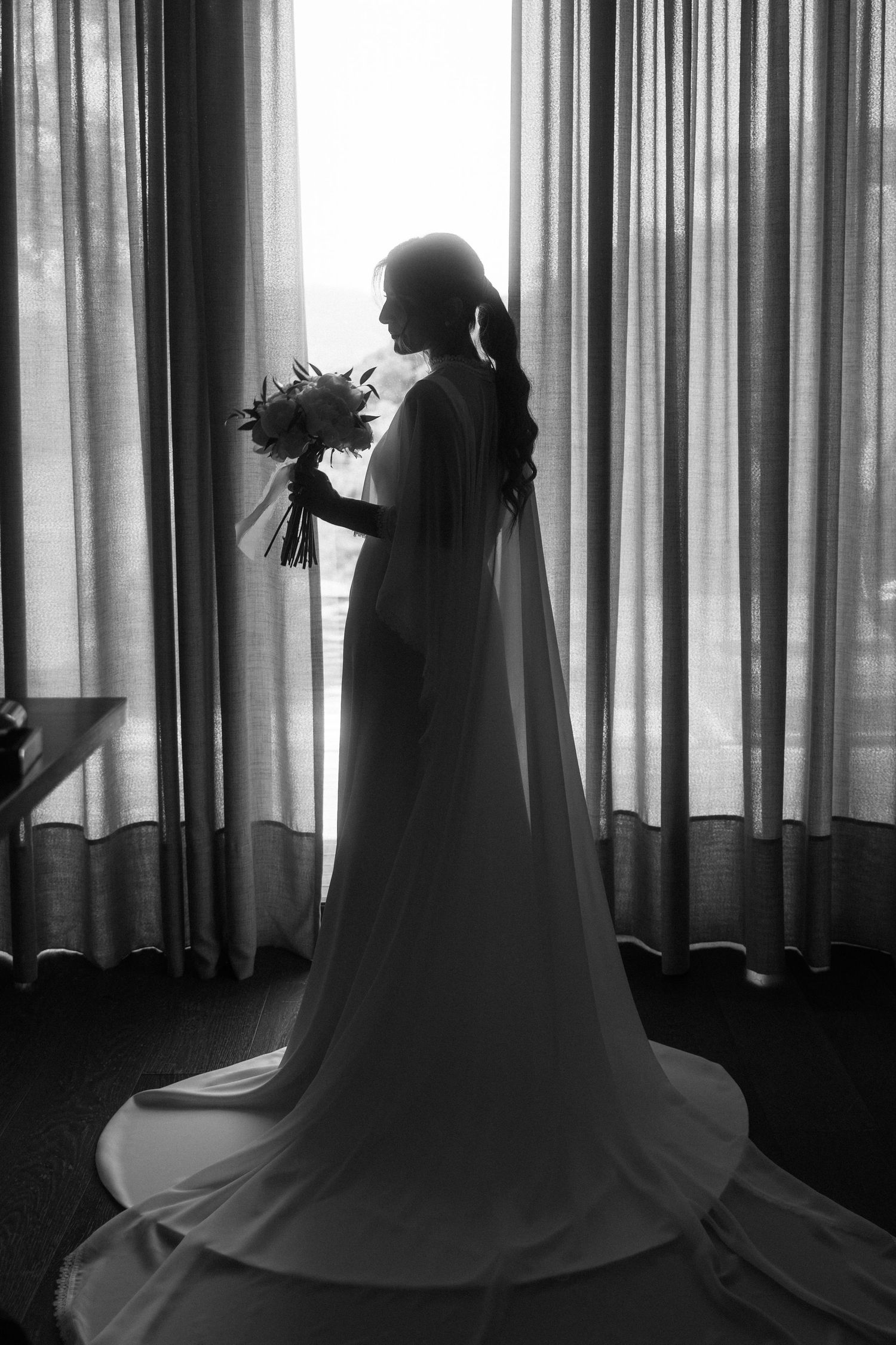 Silhouette of bride in flowing gown holding bouquet standing before large curtained window.