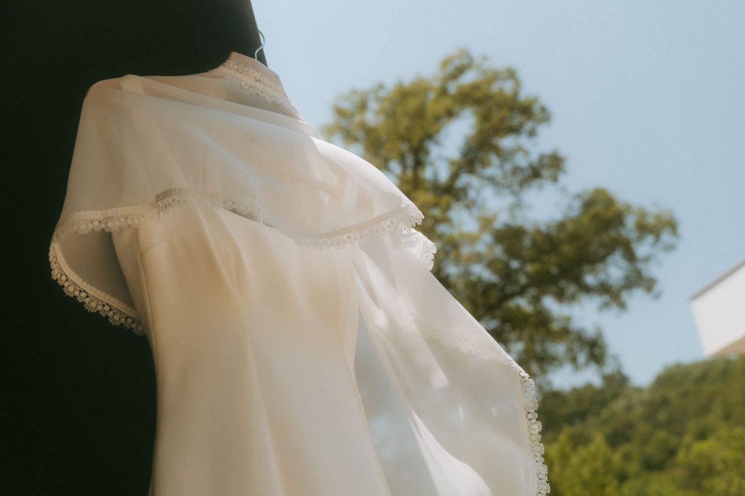 Close-up of flowing white wedding dress cape with lace trim against blue sky.