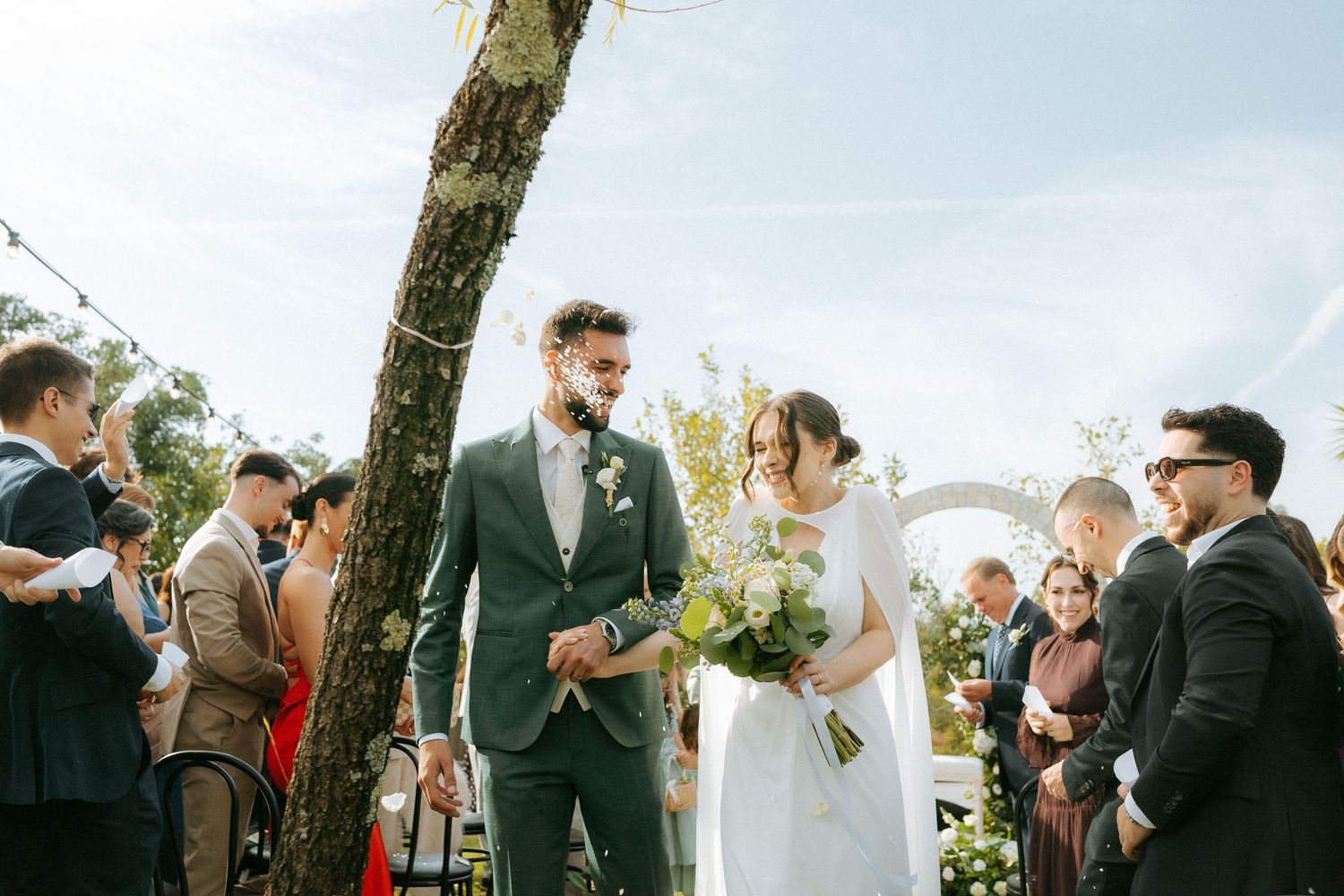 Newlyweds walk down the aisle together outdoors, bride holding a lush bouquet of white and green flowers.
