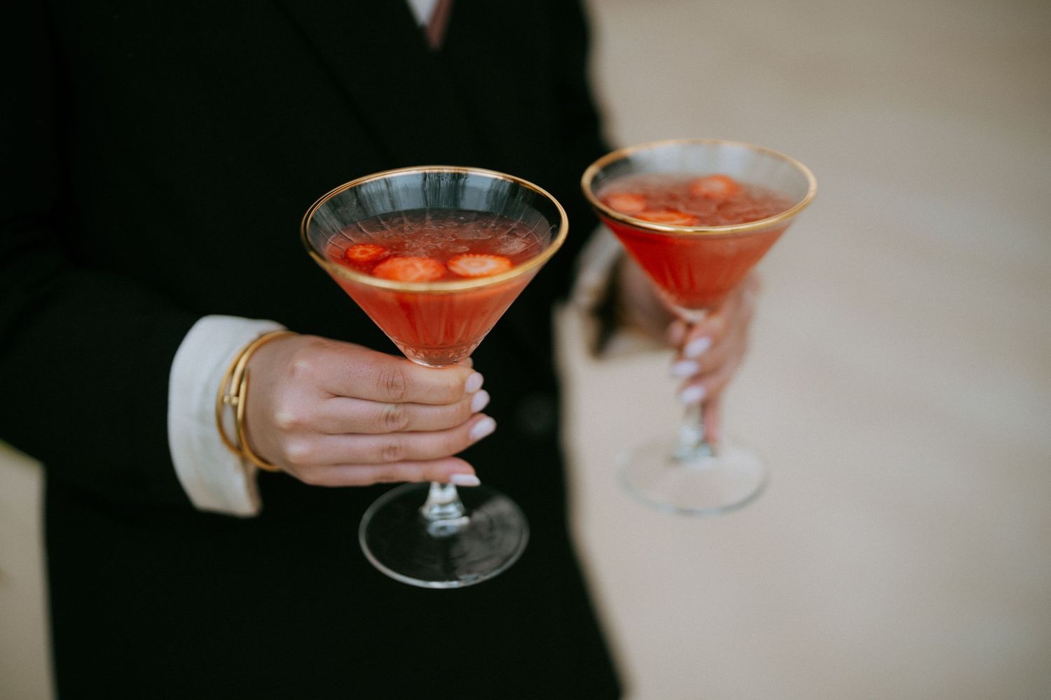 Elegant hand holding two red cocktails in gold-rimmed martini glasses at a wedding reception.