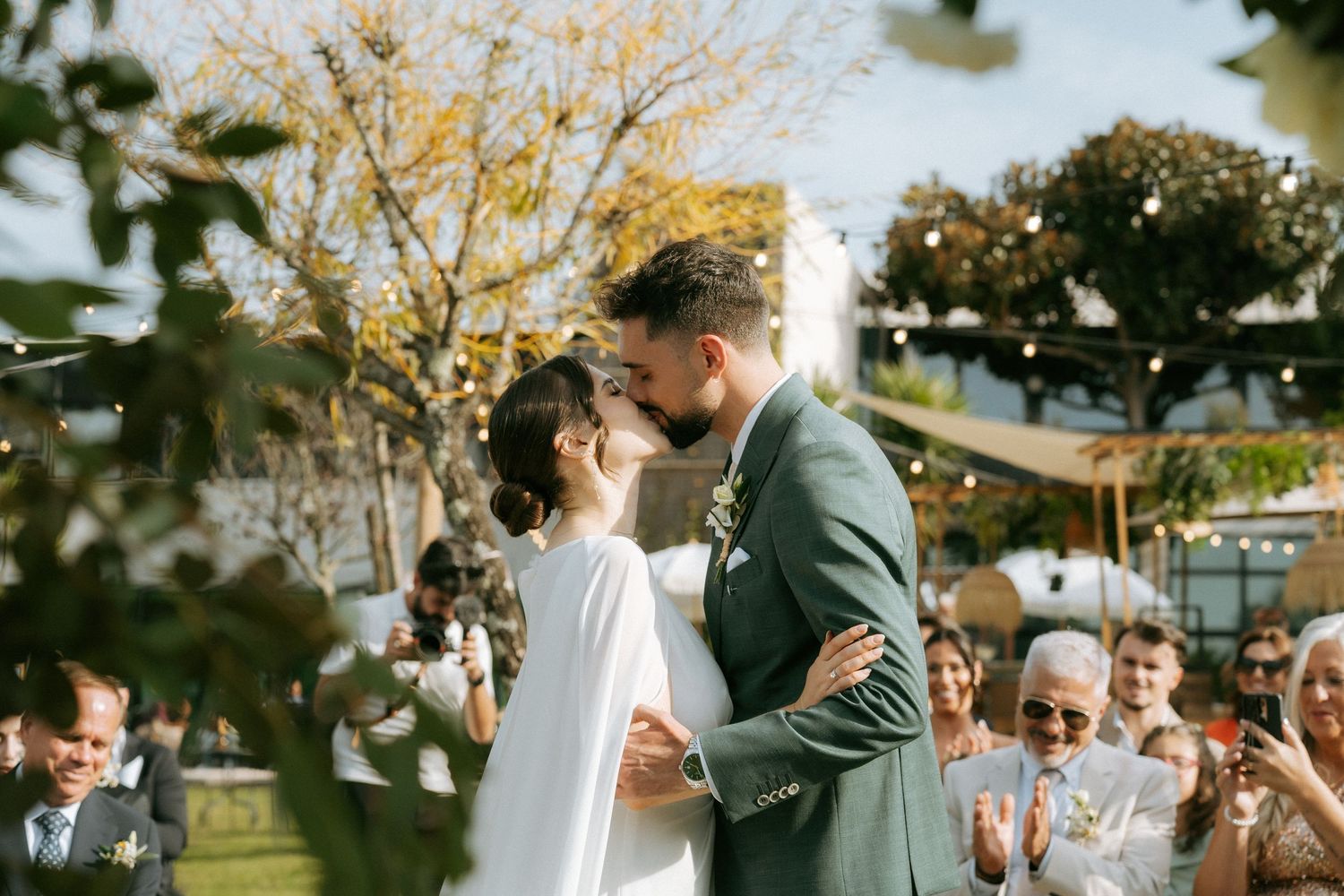 Bride and groom share their first kiss at an outdoor wedding ceremony surrounded by cheering guests.