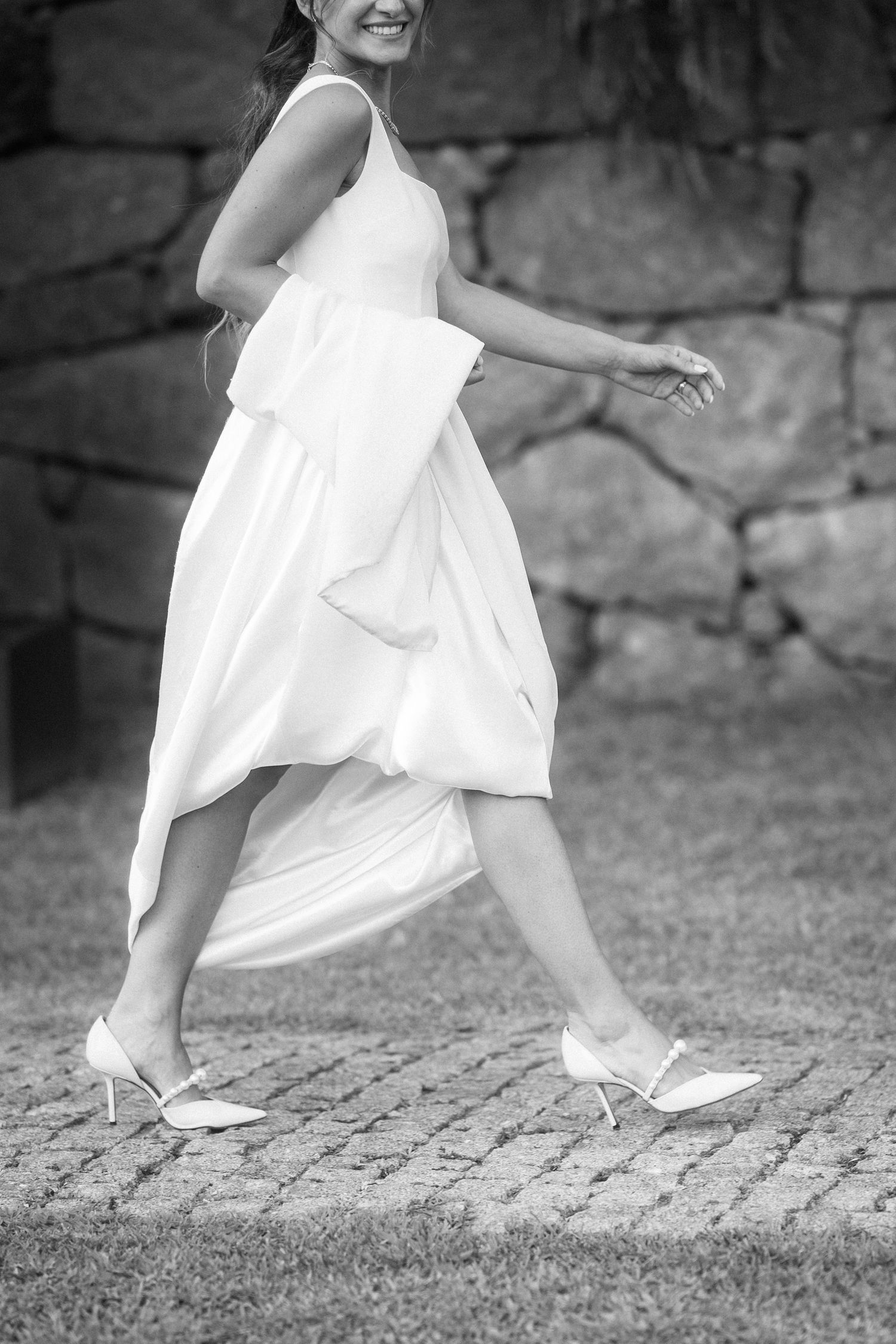 Black and white photo of a bride in a flowing white dress and heels walking outdoors.