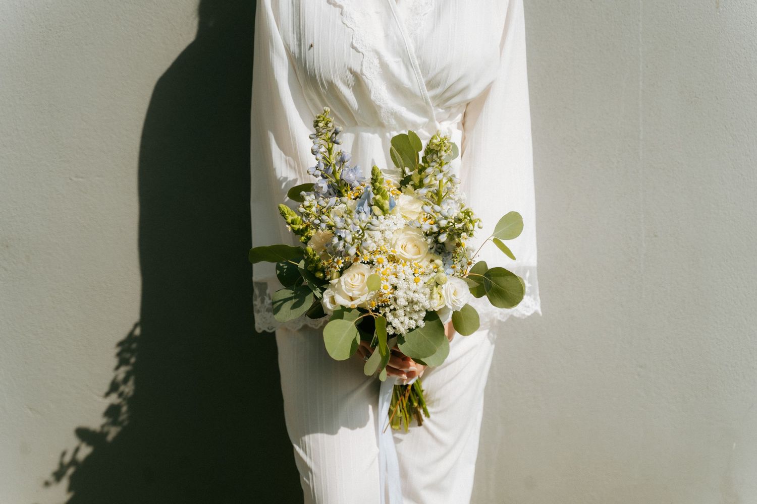 Bride in white jumpsuit holding elegant bouquet of white roses and blue wildflowers against white wall.