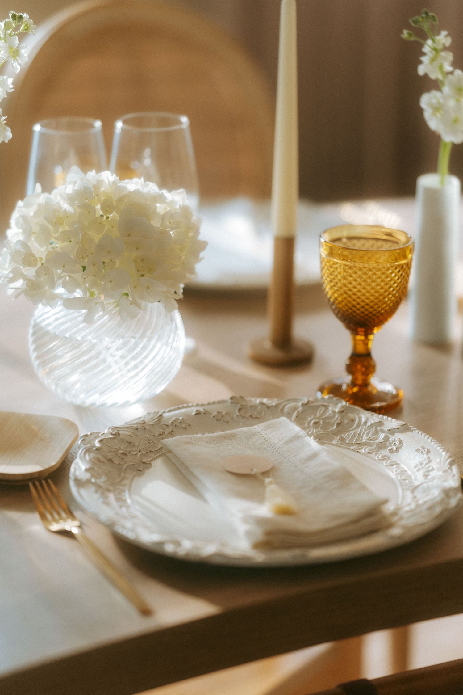 Close-up of a wedding place setting with ornate white plate, linen napkin, gold cutlery, and amber goblet.