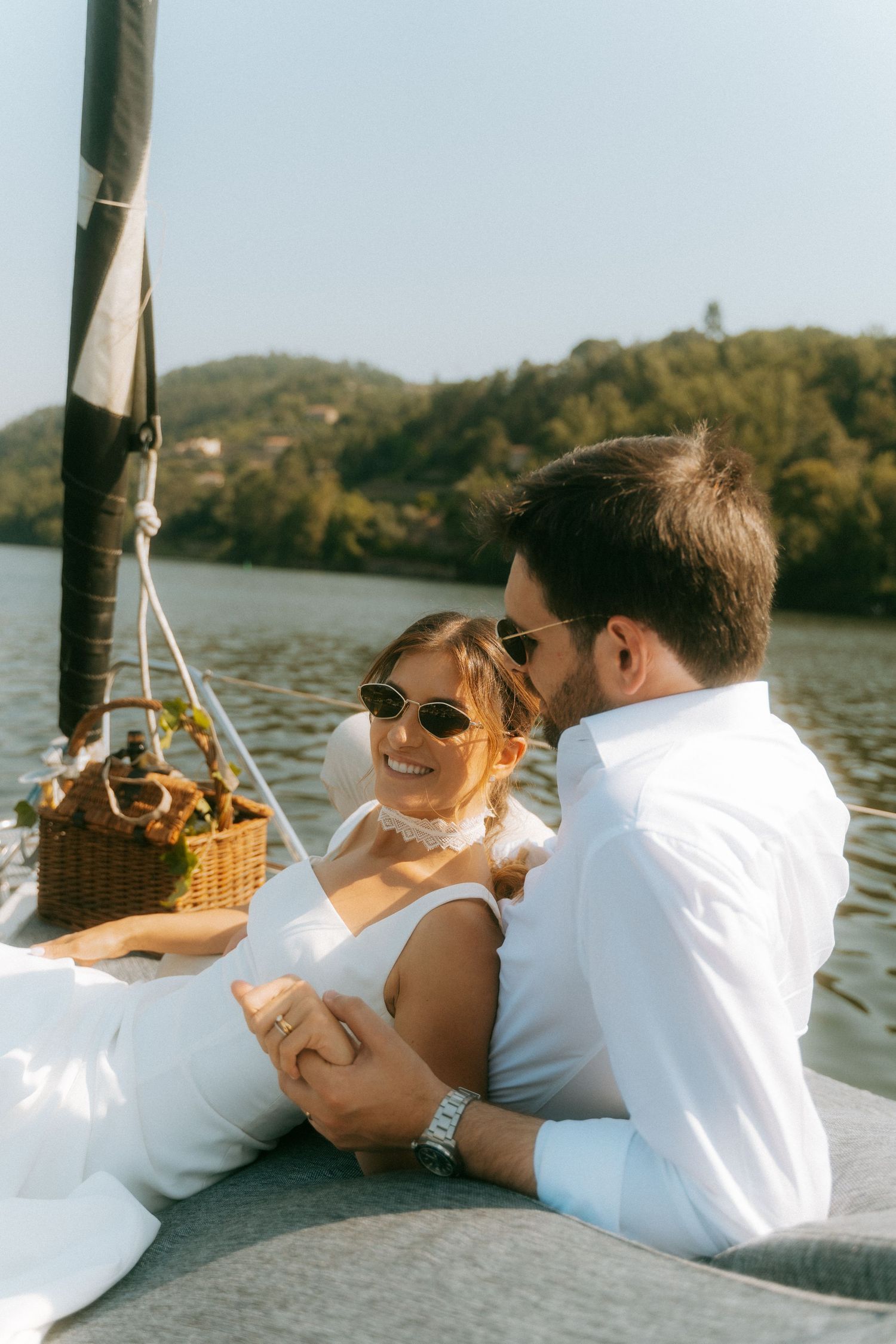 Smiling bride and groom in white attire sitting together on a sailboat with scenic hills behind.