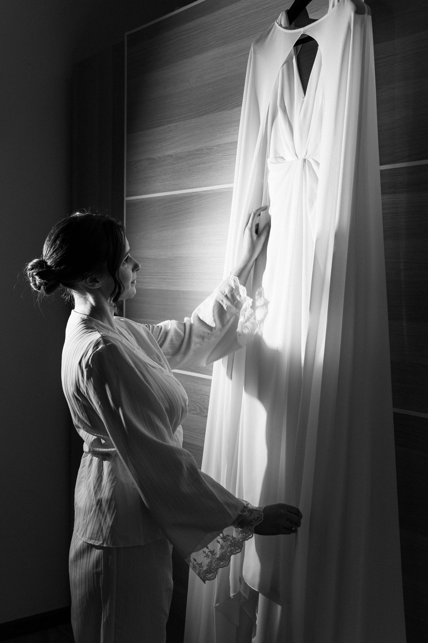 Black and white photo of bride in robe reaching for white wedding dress hanging on wardrobe.