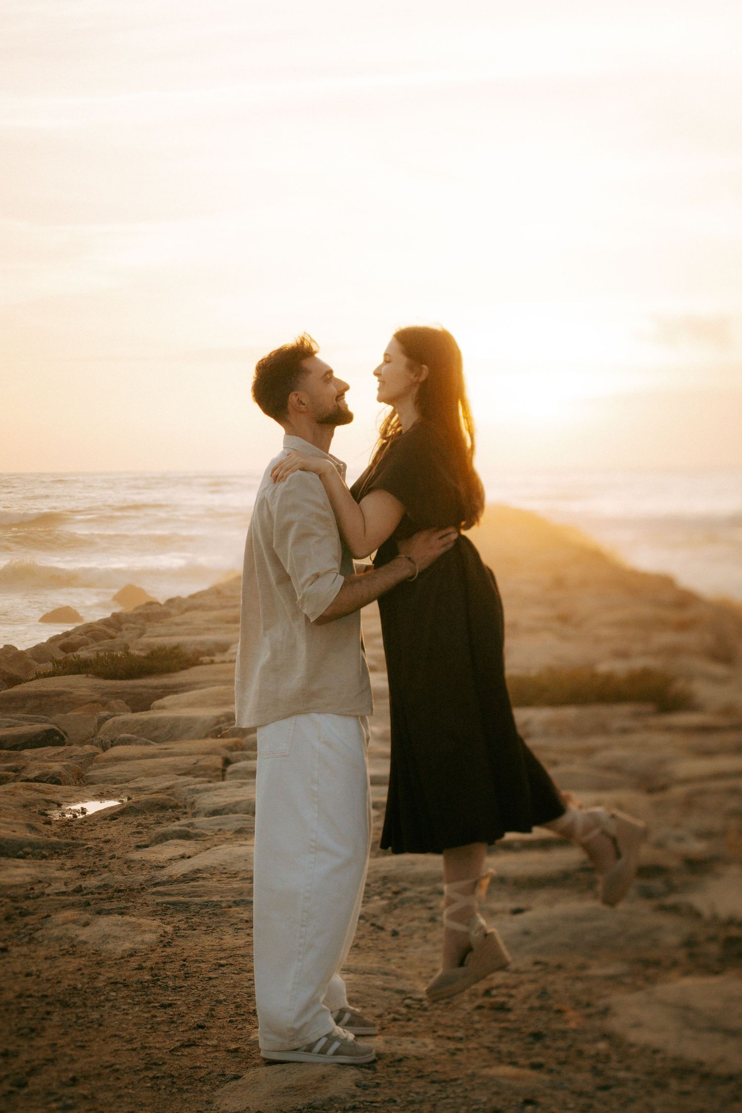 Couple embracing romantically on rocky beach at golden sunset, man in white pants and woman in black dress.