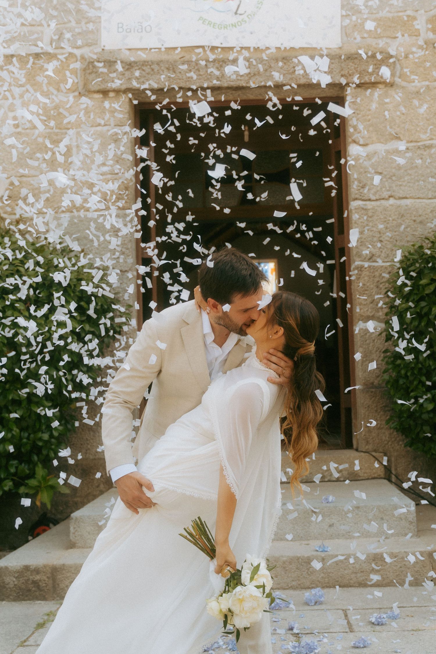 Newlyweds sharing first kiss under shower of white confetti outside stone church entrance.