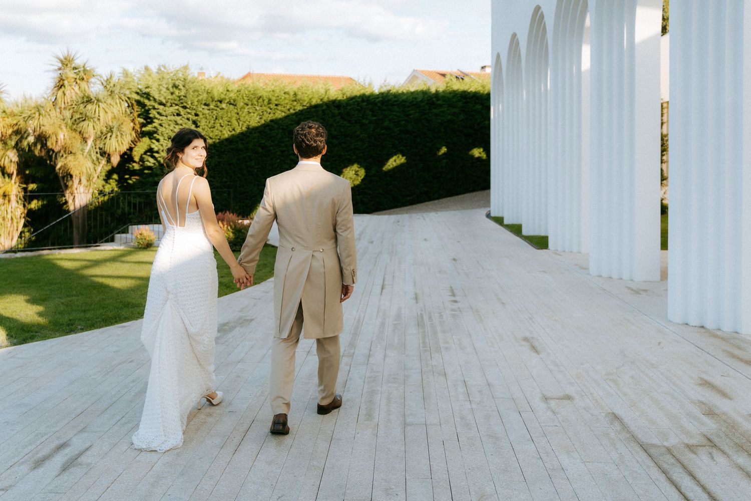 Bride and groom walk hand in hand along a white columned outdoor wedding venue pathway.