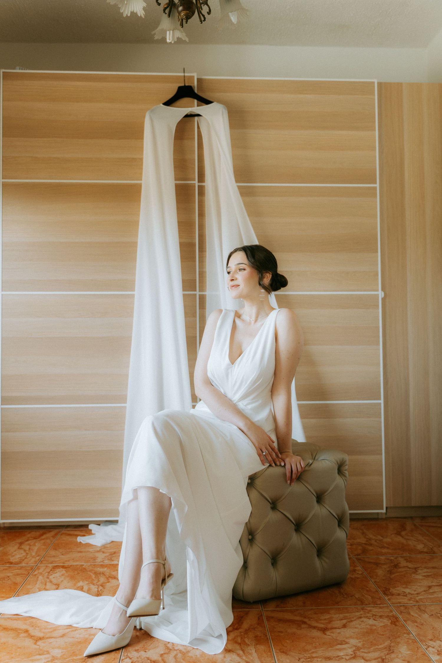 Elegant bride in white gown sitting on tufted ottoman beside hanging wedding dress in bright room.
