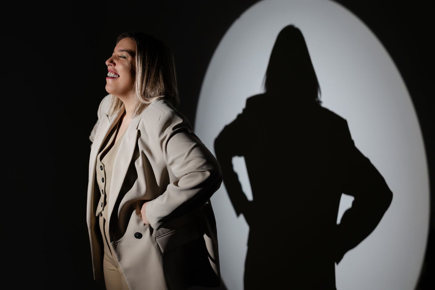 Woman in beige trench coat poses dramatically with bold circular light and shadow backdrop.