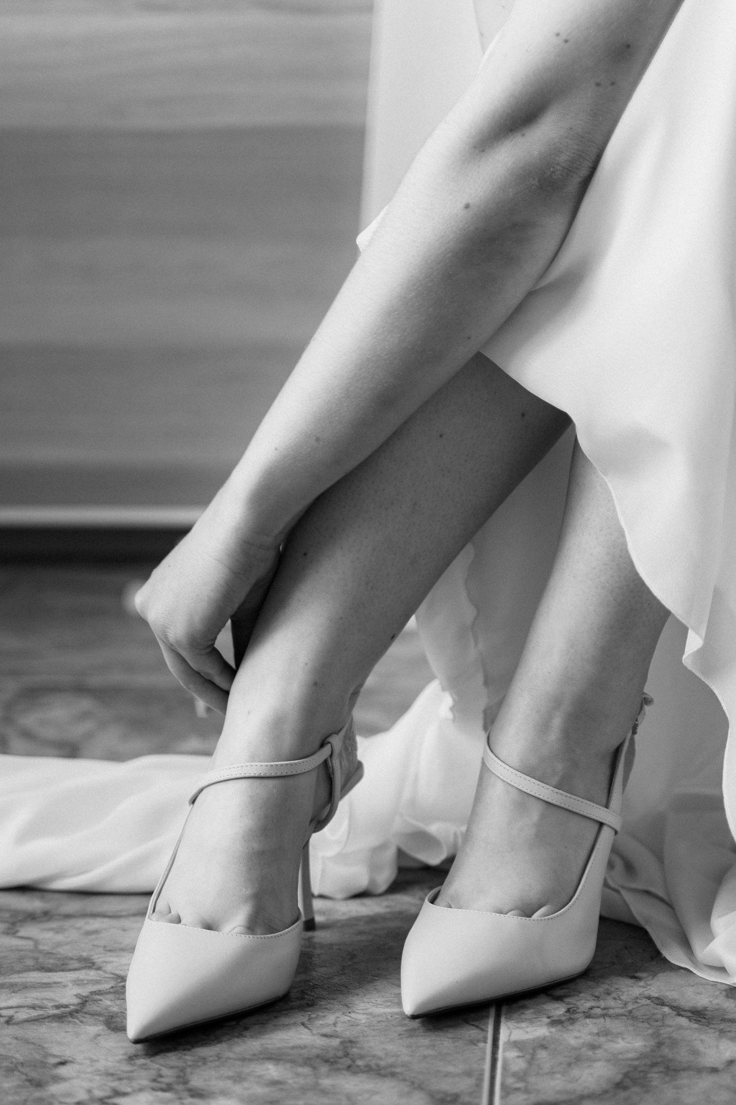 Black and white close-up of bride fastening strappy pointed-toe heels beneath white wedding dress.