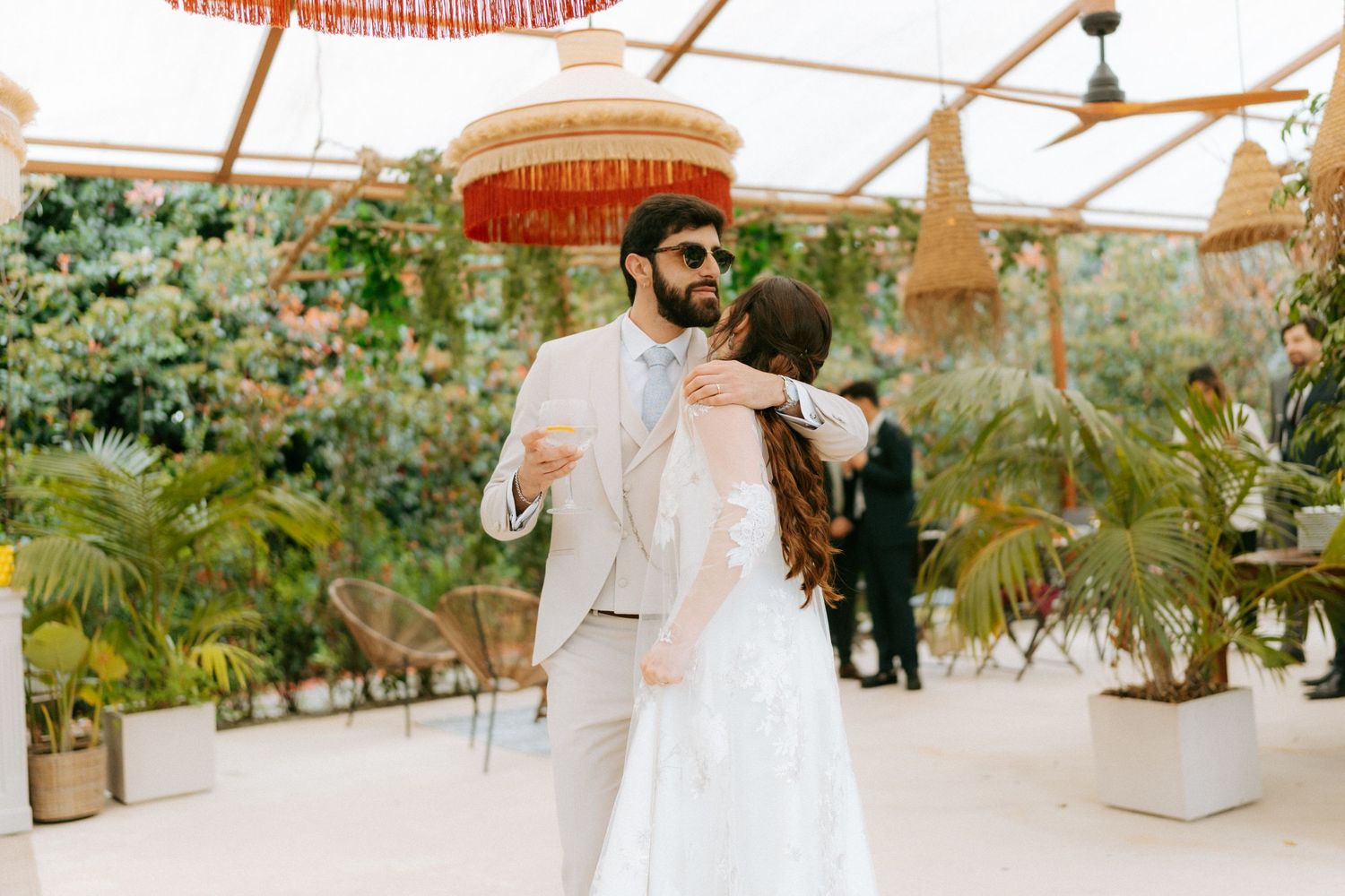 Groom in beige suit and sunglasses shares first dance with bride in a tropical greenhouse venue.