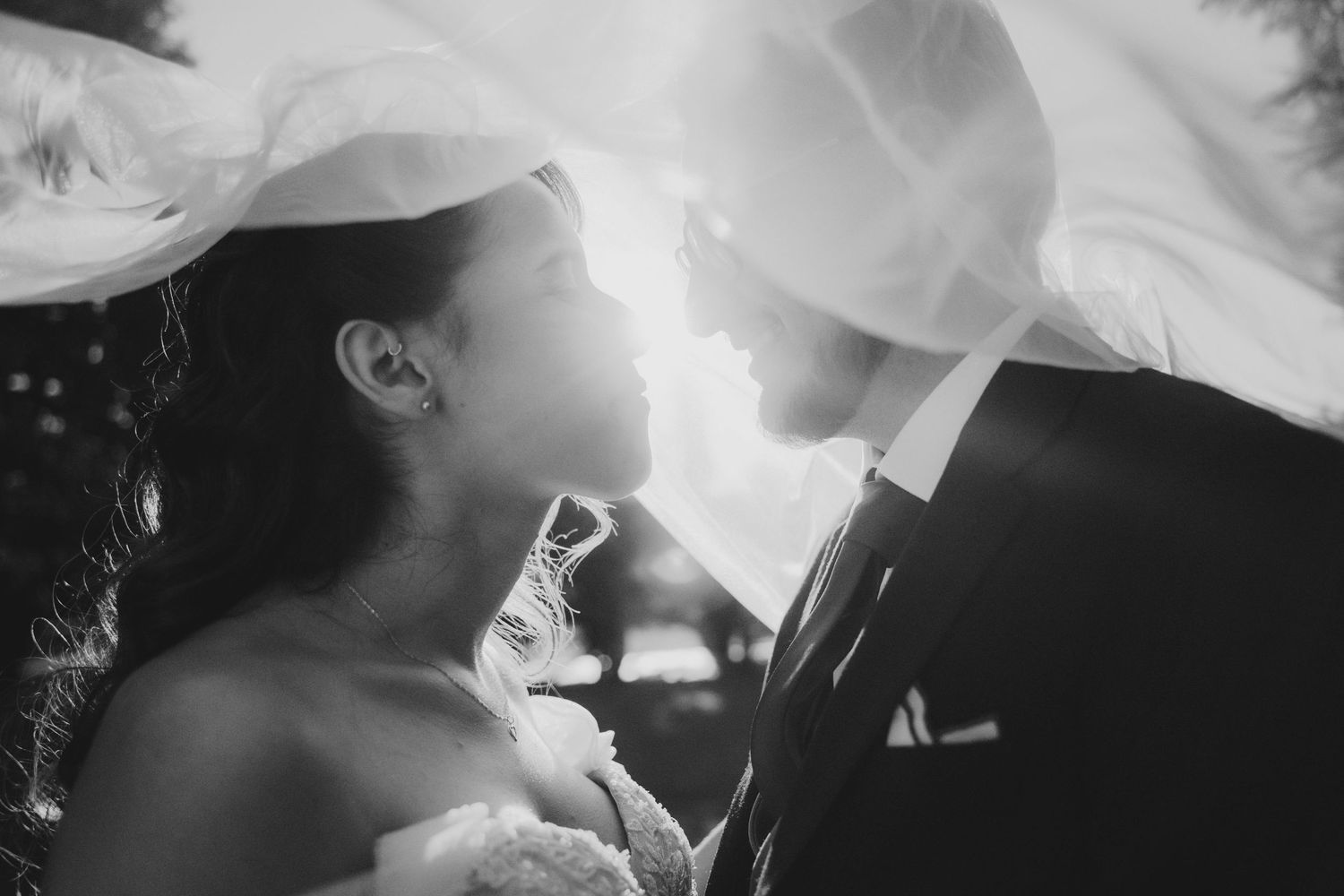 Black and white photo of bride and groom gazing at each other under a flowing veil with sunlight.