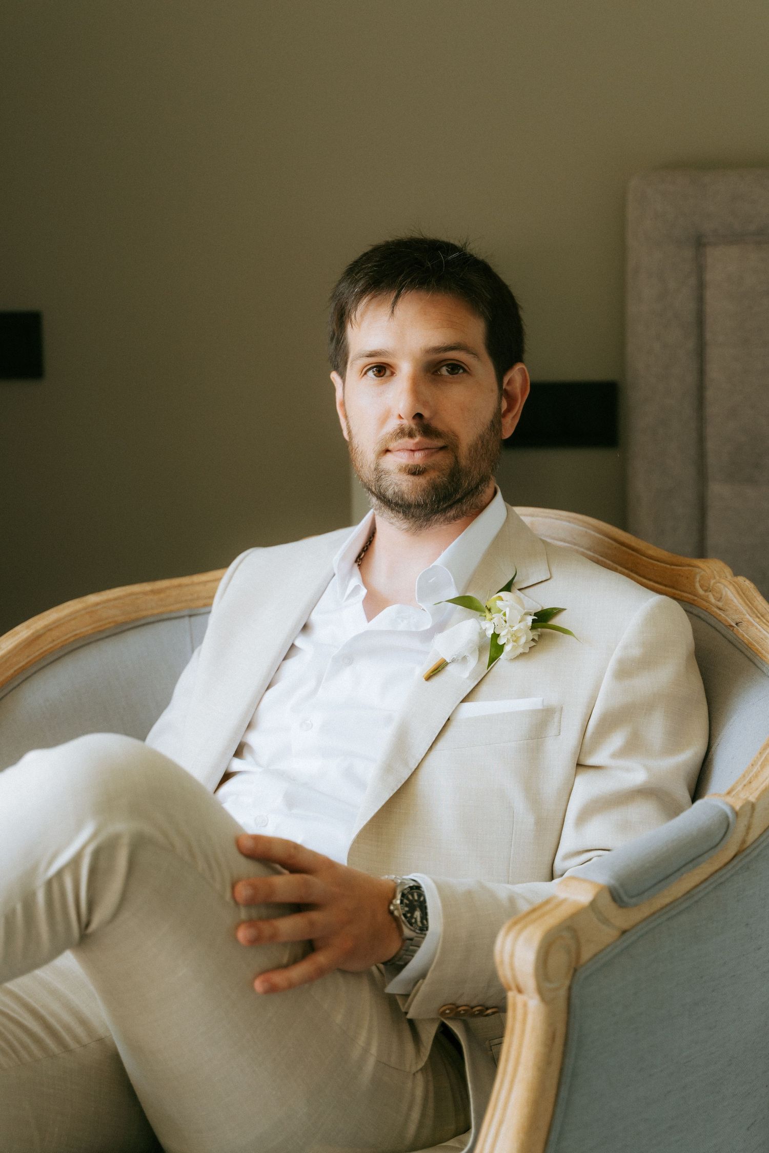 Groom in beige suit with white boutonniere seated in elegant chair at wedding venue.