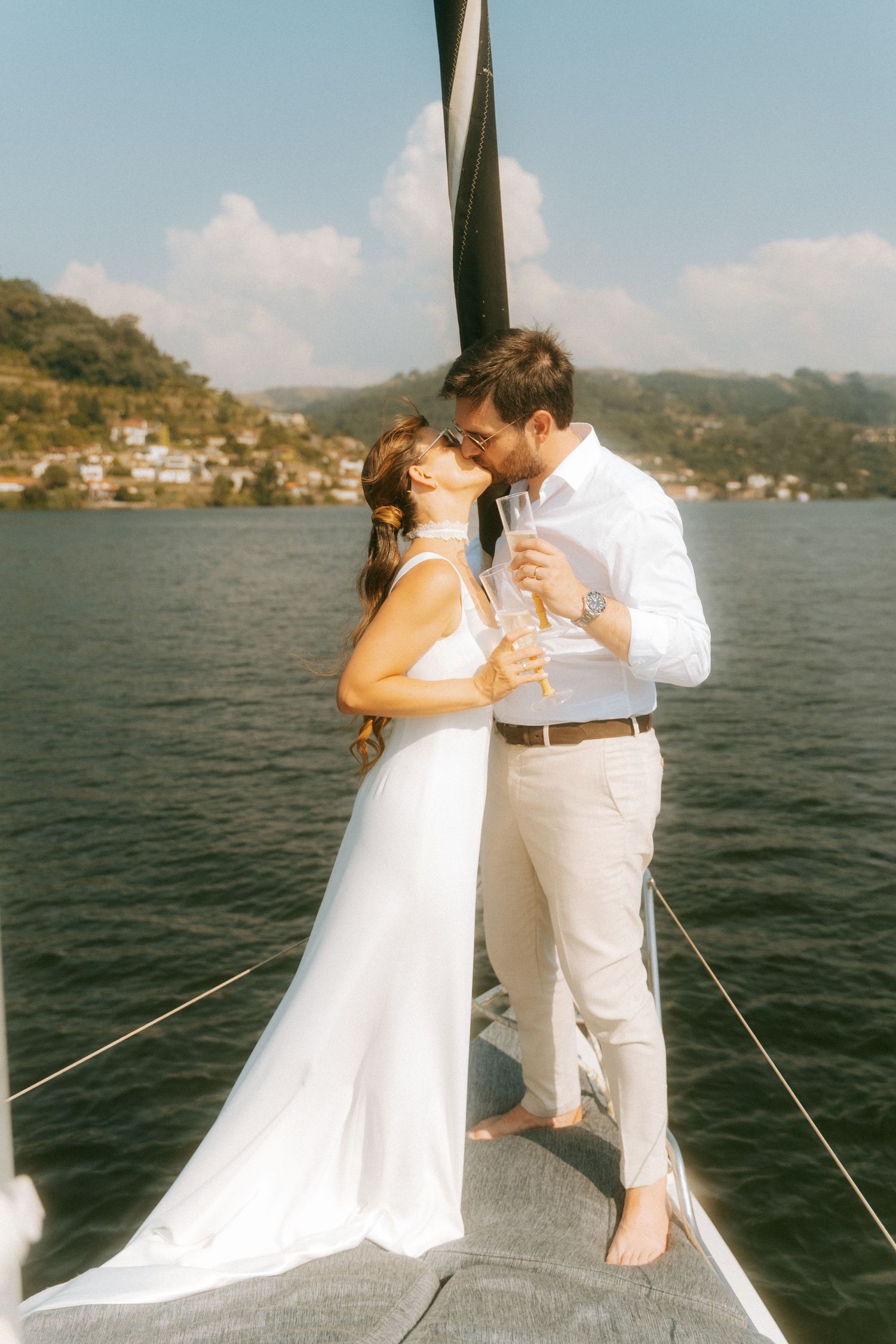 Bride and groom kissing while holding champagne glasses on a sailboat with blue sky and hills.