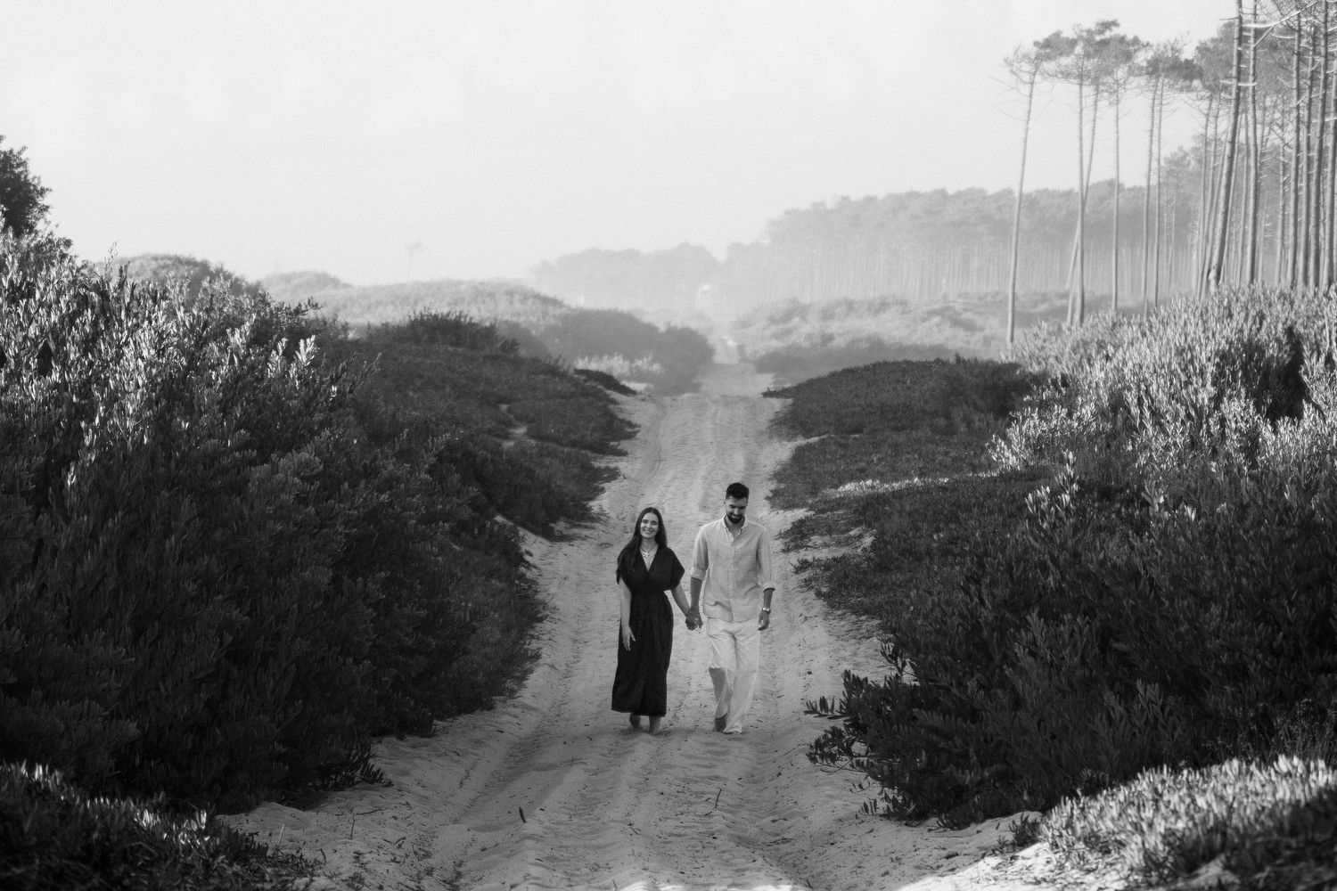 Bride and groom walk hand in hand down a misty dirt path surrounded by trees in black and white.