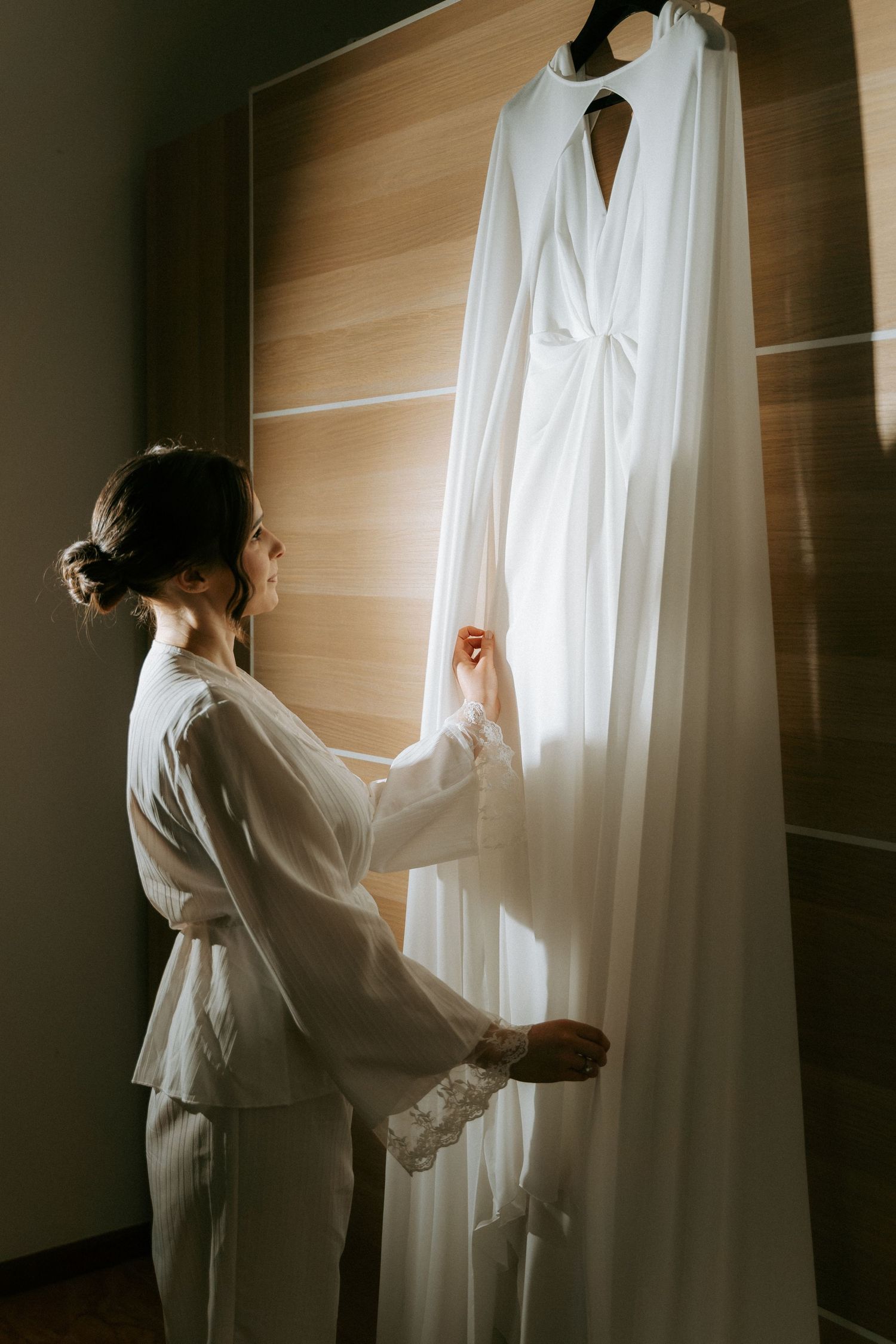 Bride in white robe admiring her wedding dress hanging on a wooden door in soft natural light.