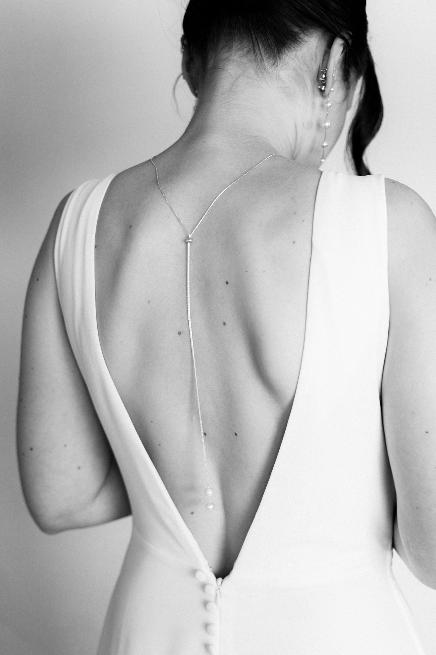 Black and white photo of a bride in a backless white gown with a delicate back necklace, showcasing elegant bridal style.