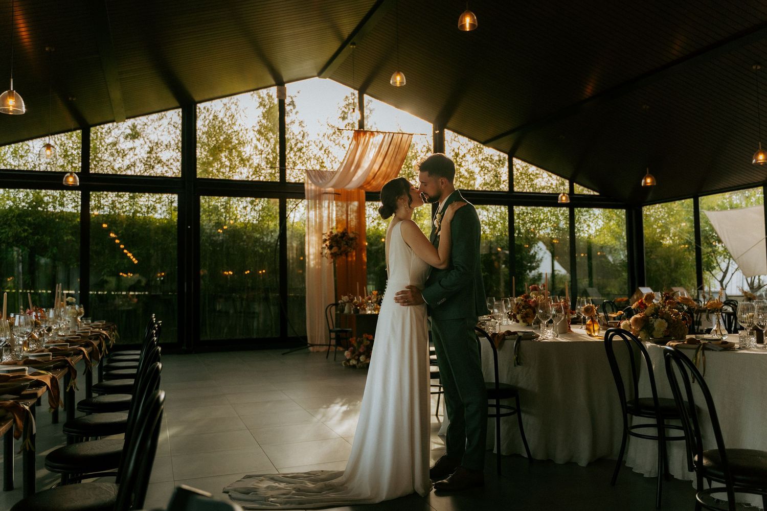 Bride and groom share a romantic moment in a sunlit glass pavilion wedding reception venue.