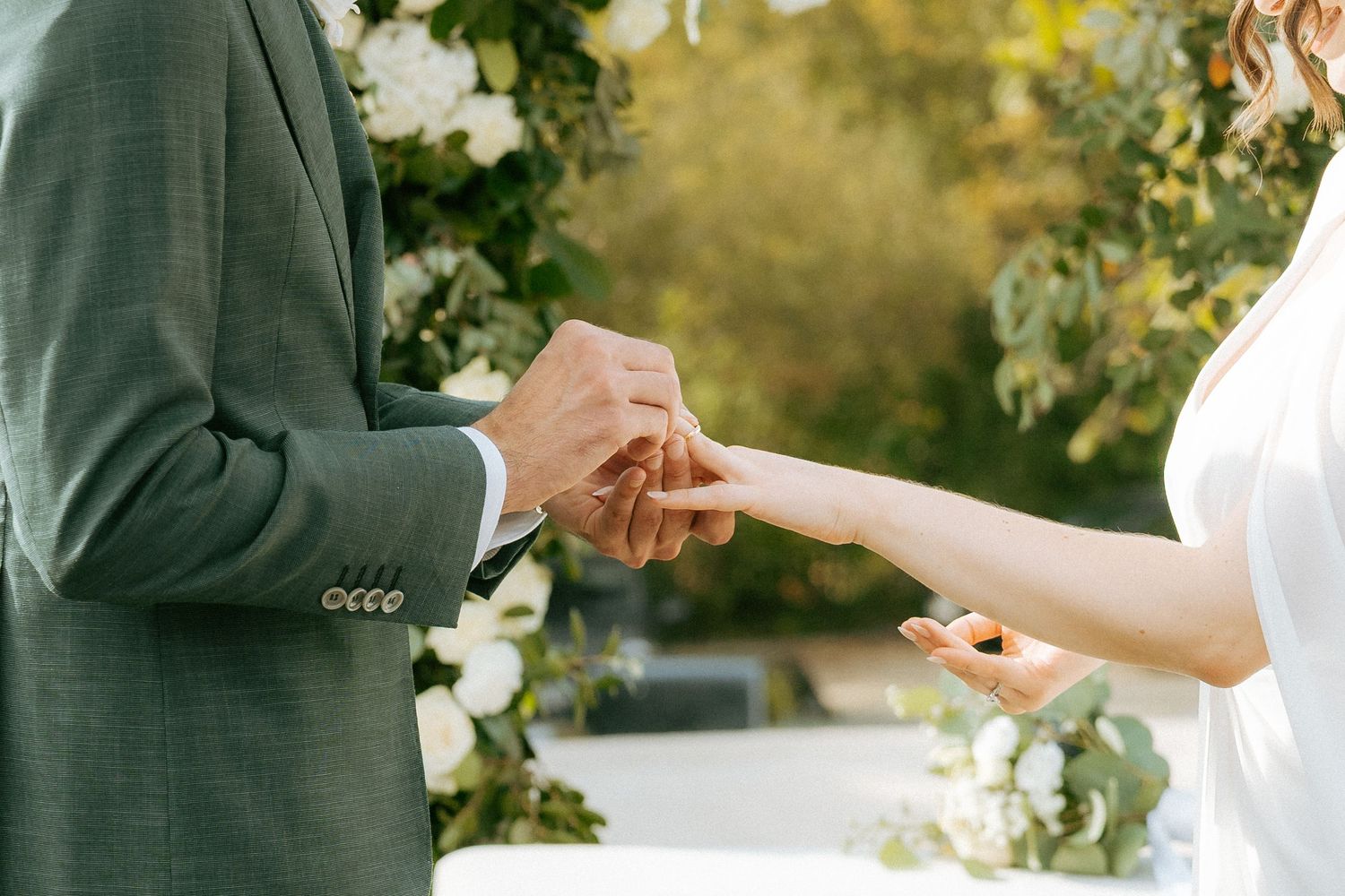Groom placing wedding ring on bride's finger during outdoor ceremony with white floral arch backdrop.