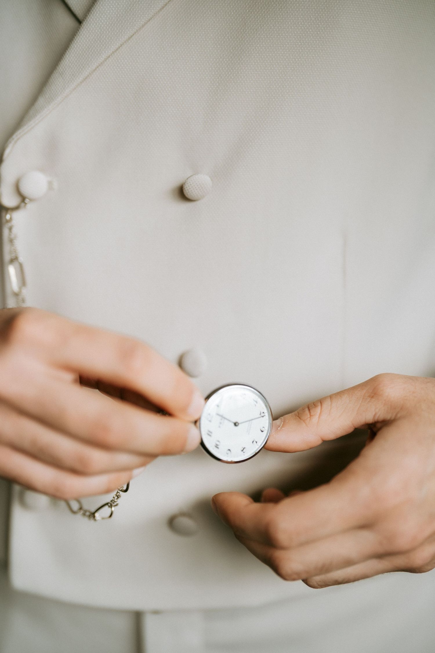 Groom in beige suit holding a vintage silver pocket watch with chain against his jacket buttons.