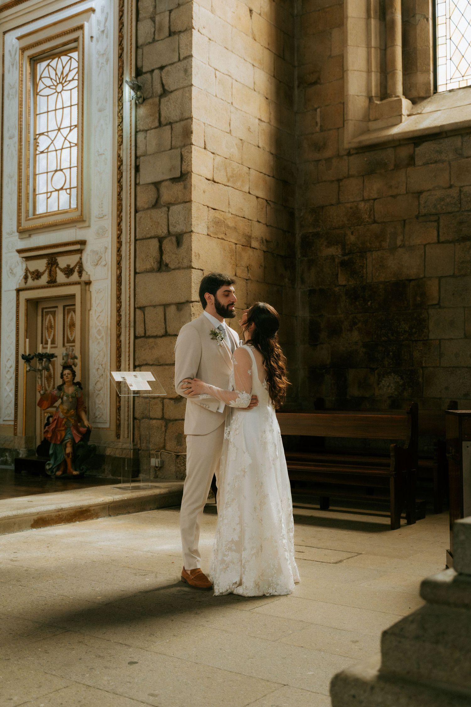 Bride and groom share a romantic embrace inside a sunlit historic stone church after their wedding.