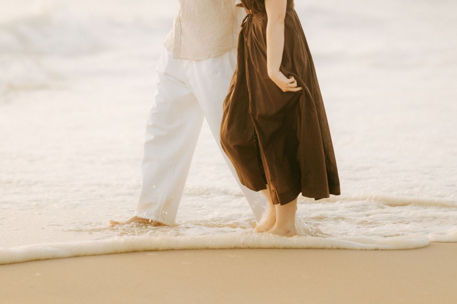 Barefoot couple walking together in ocean waves on sandy beach, woman in flowing brown dress.